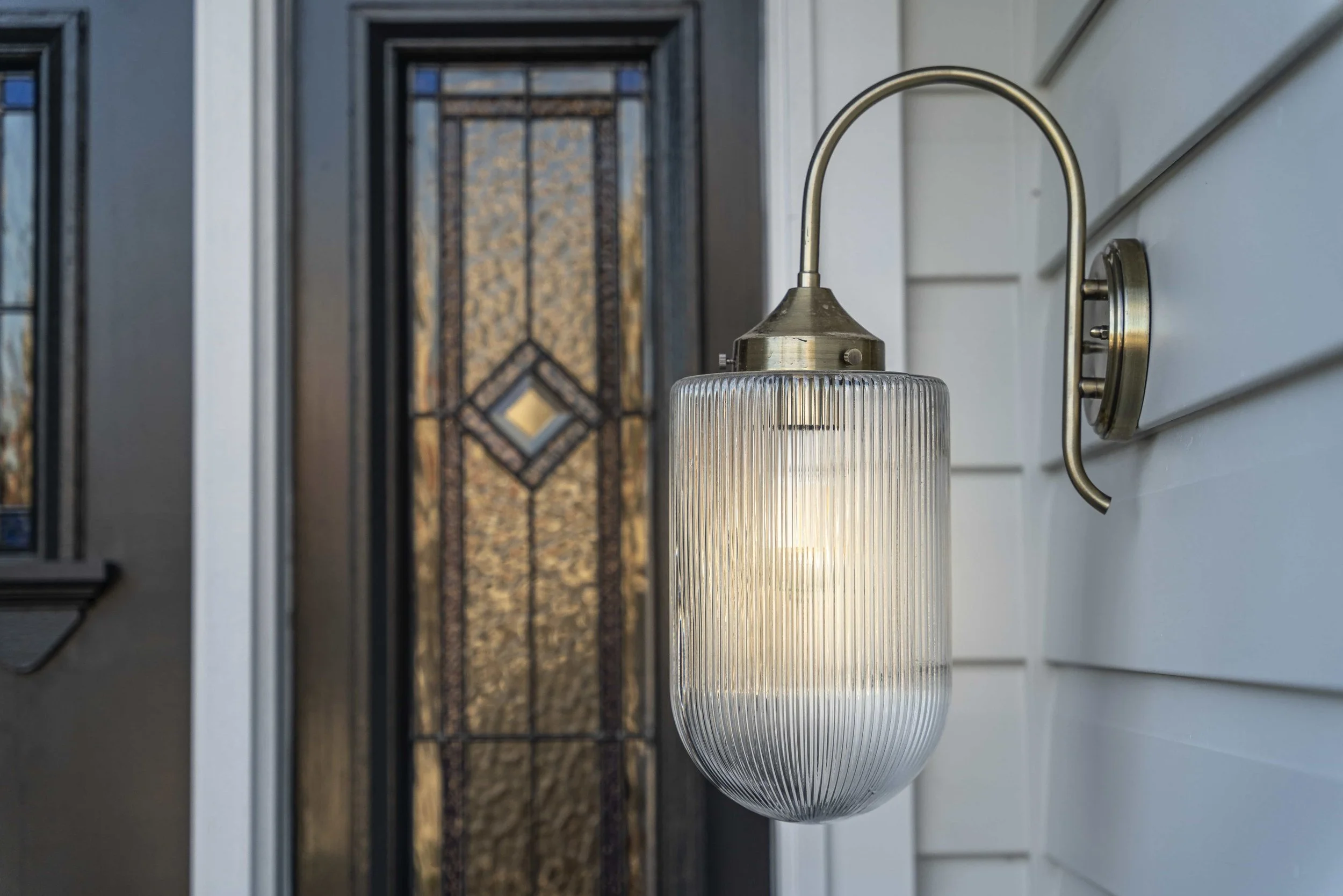A wall-mounted outdoor light fixture with a ribbed glass shade on a white house exterior, with a decorative door in the background.