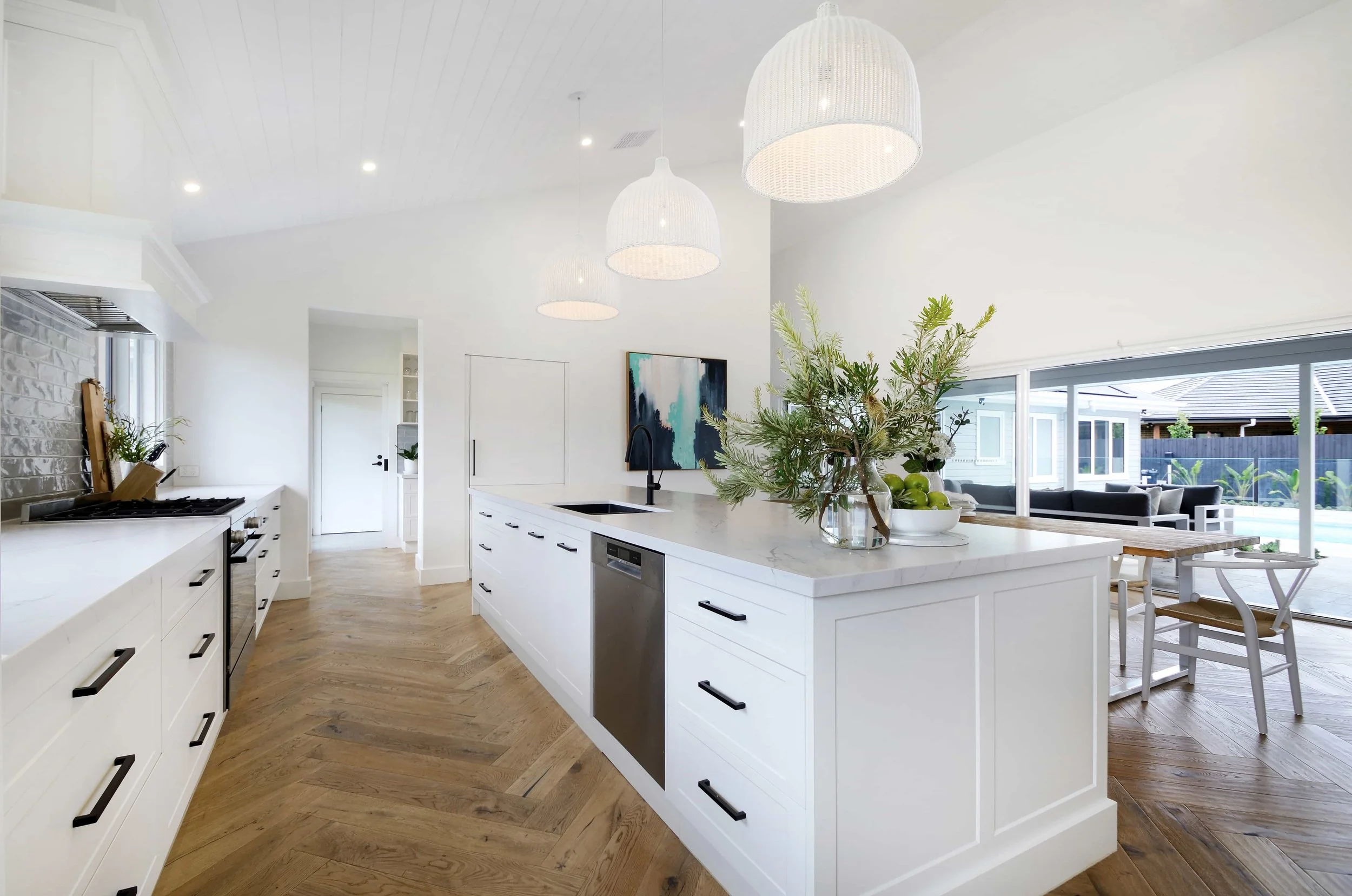 Modern kitchen with routed white cabinet doors and island, timber herringbone floor, large window facing backyard, and hanging white pendant lights over the island. The ceiling over the kitchen is lined with VJ board.