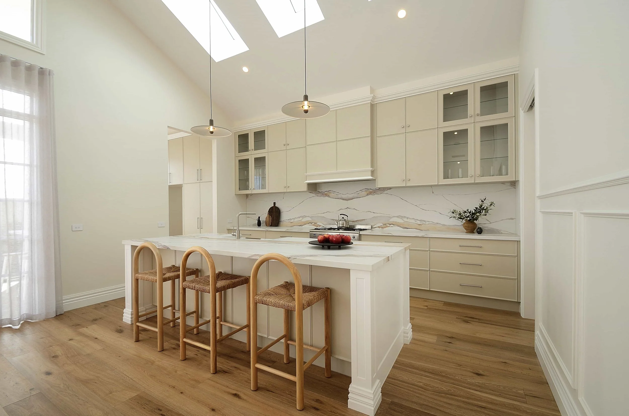 Bright kitchen with white cabinetry, a marble splashback, an island with a marble benchtop and traditional detailing. Skylights and pendant lights illuminate the space, with a potted plant and apples on the counter.