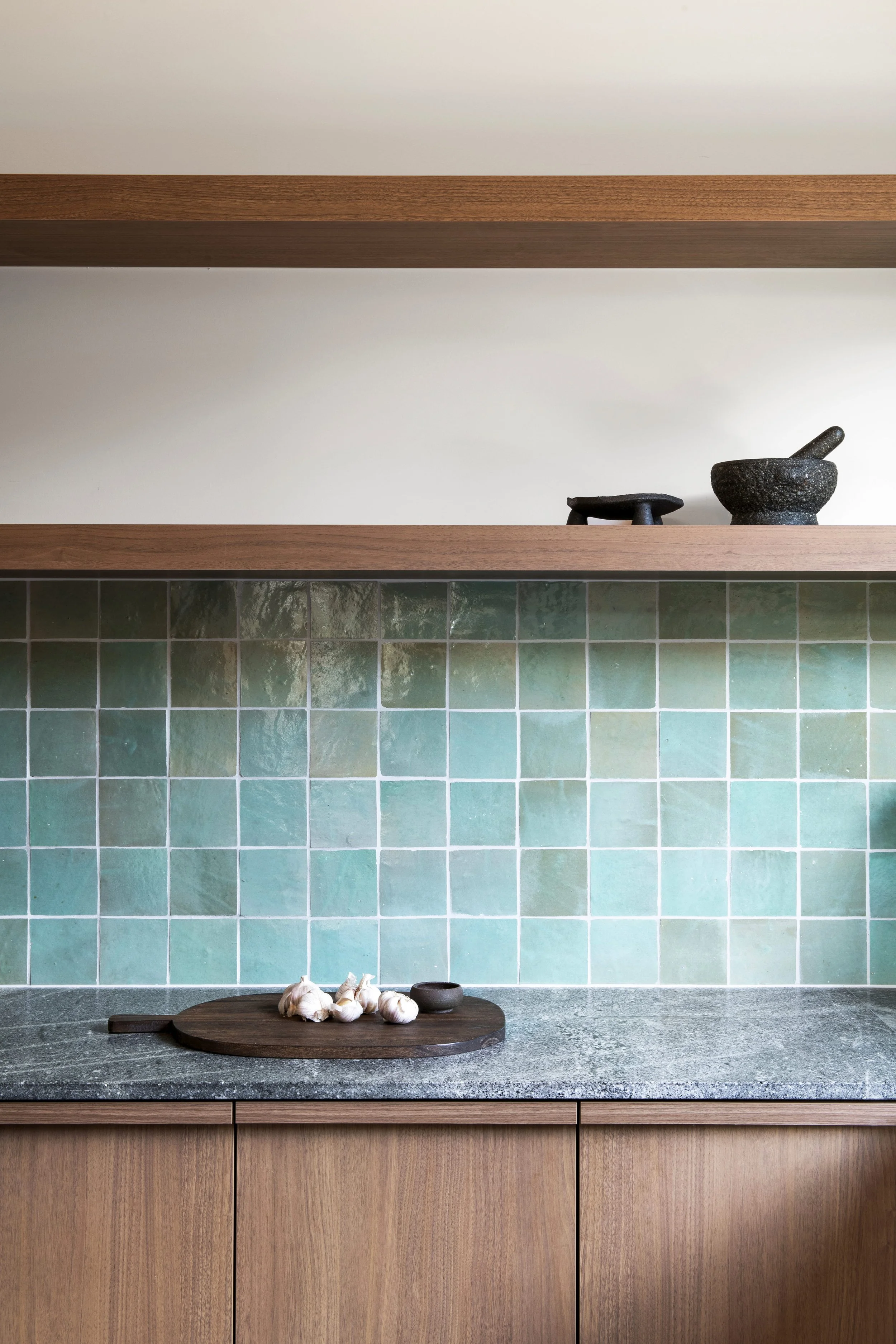 Kitchen with green tiled backsplash, timber cabinets, grey stone benchtop, and floating shelves with a mortar and pestle.