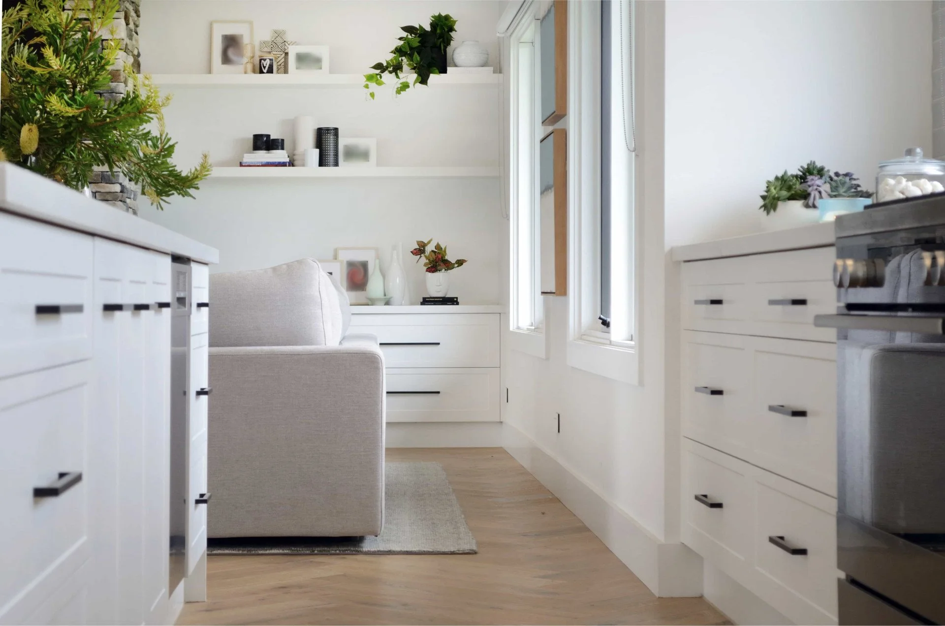 Bright white kitchen with natural light, decorated with green plants, framed art, and neutral-colored furniture.