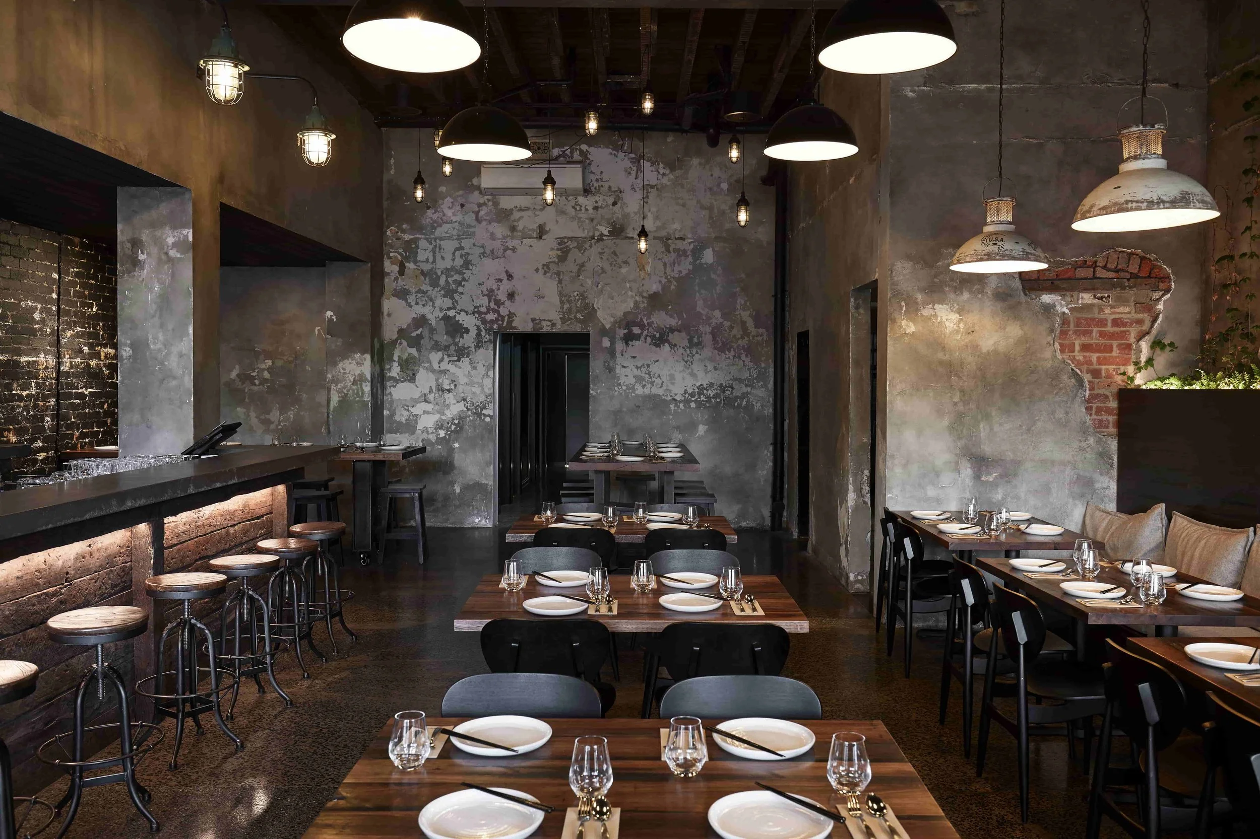 Interior of a rustic restaurant with exposed brick and distressed walls, pendant and industrial-style ceiling lights, and tables set with plates, glasses, and cutlery.