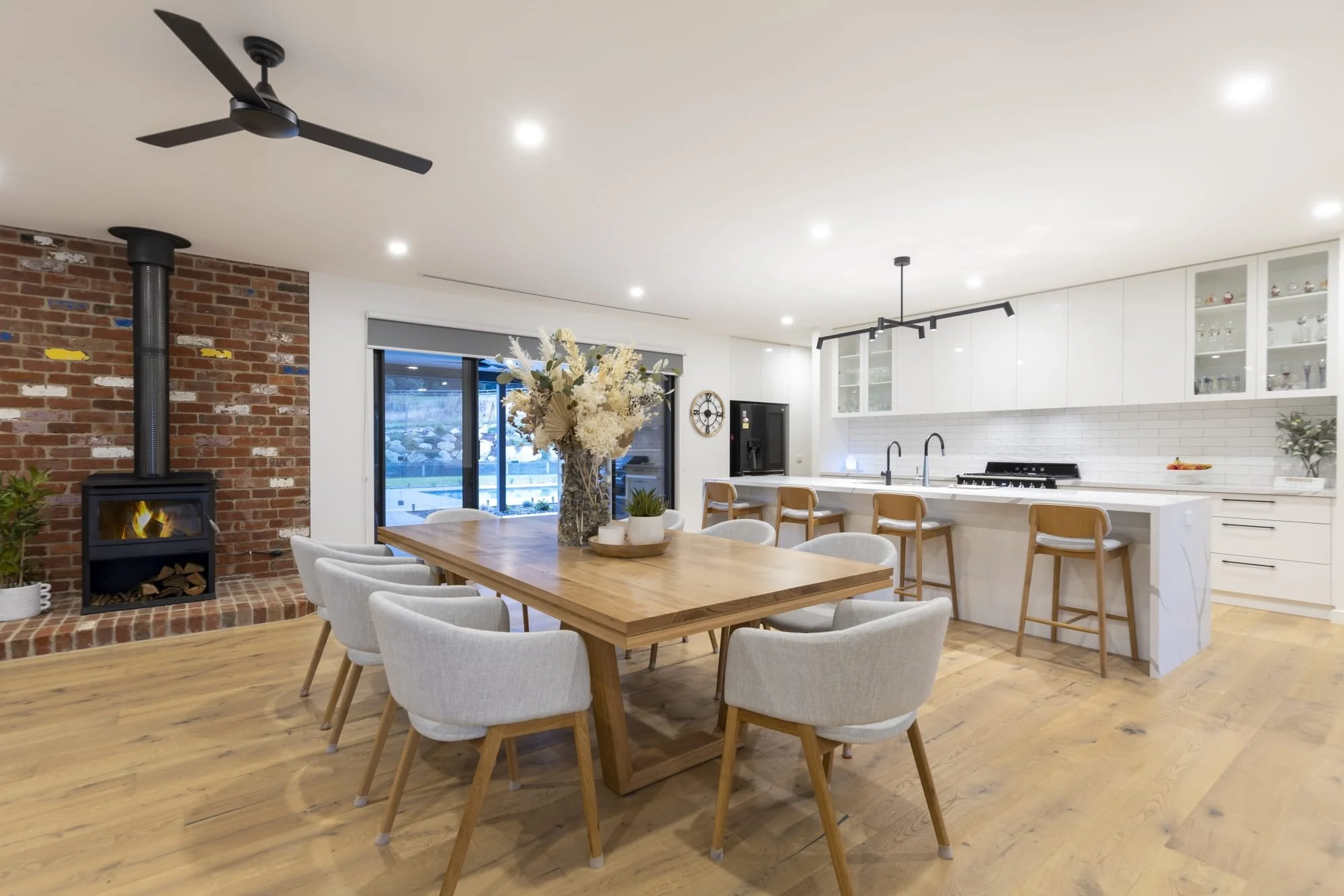 Modern kitchen and dining area with a wooden table surrounded by white upholstered chairs, a large floral centerpiece, a brick fireplace with a wood stove, white cabinets, and a sliding glass door leading outside.