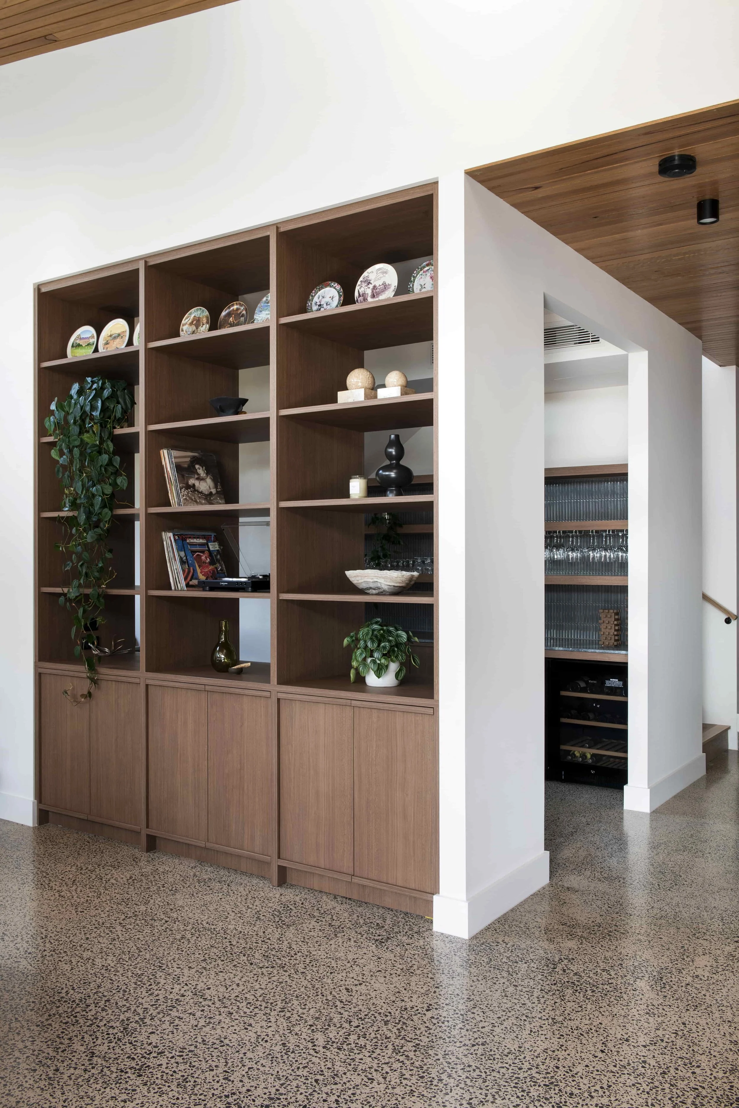 Timber built-in cabinetry bookshelf with decorative plates, books, and plants, next to a white wall opening to a room with a wine cooler and glassware.
