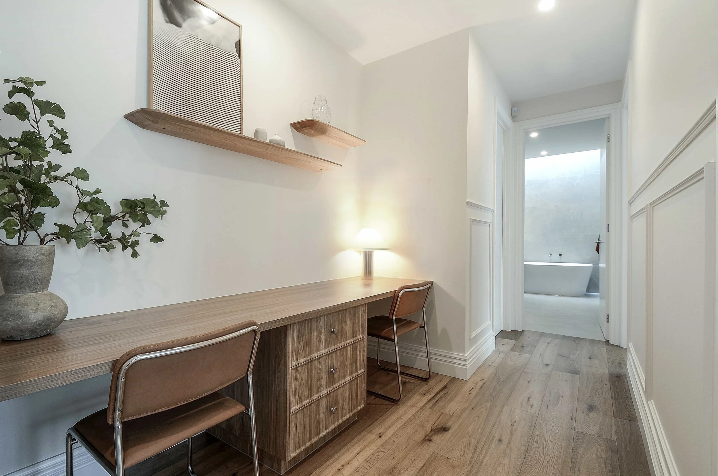 A minimalist hallway study nook with built in timber cabinetry, two brown chairs, a large potted plant, and floating wooden shelves with decorative items. A lamp provides warm lighting, and a doorway leads to a bathroom with a freestanding bathtub.