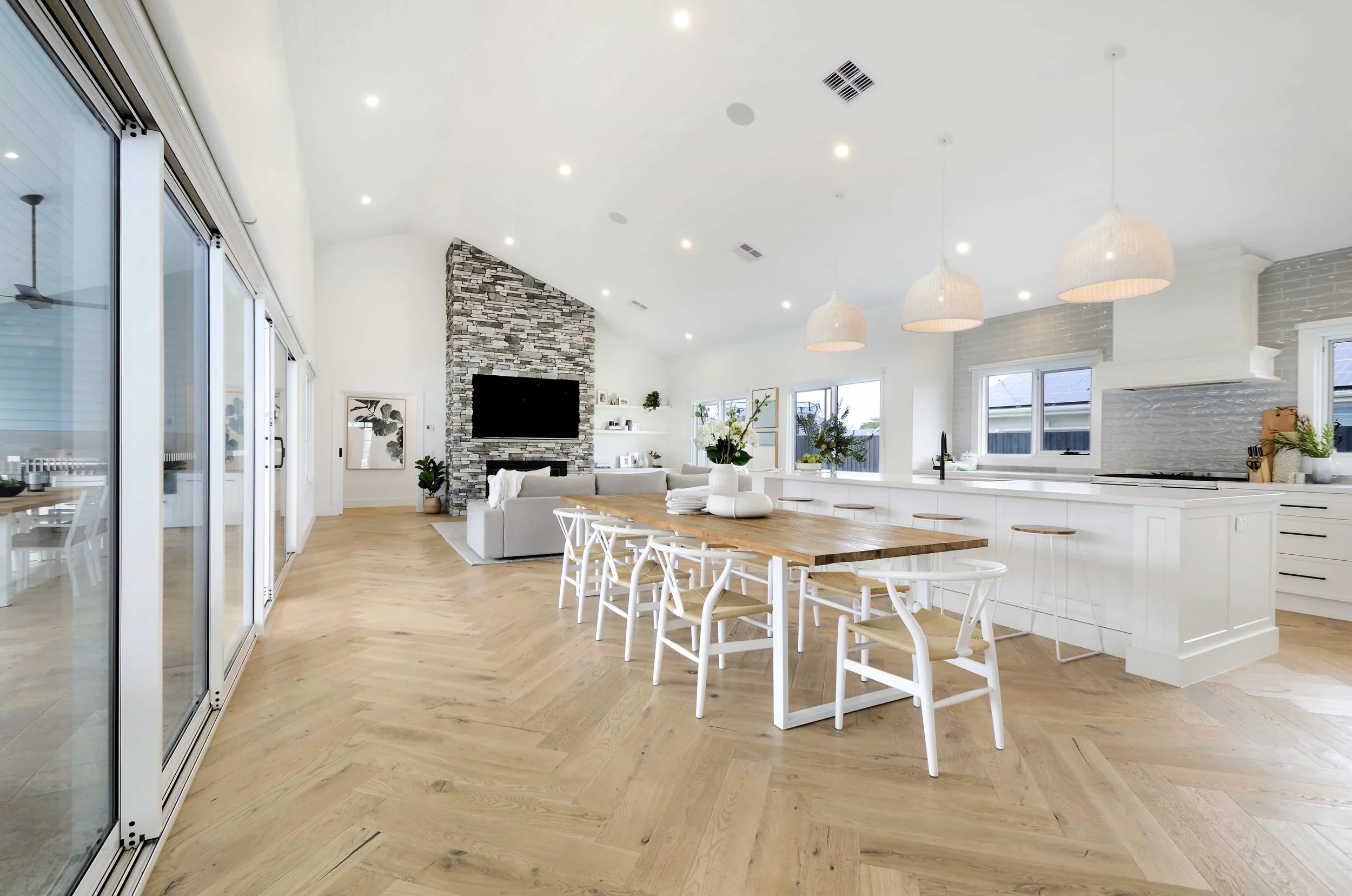 Modern open-plan living space with a dining area, a kitchen with white cabinets, a stone clad fireplace with a mounted TV, large windows, and pendant lighting. There is a cathedral ceiling with an off centre ridge line.