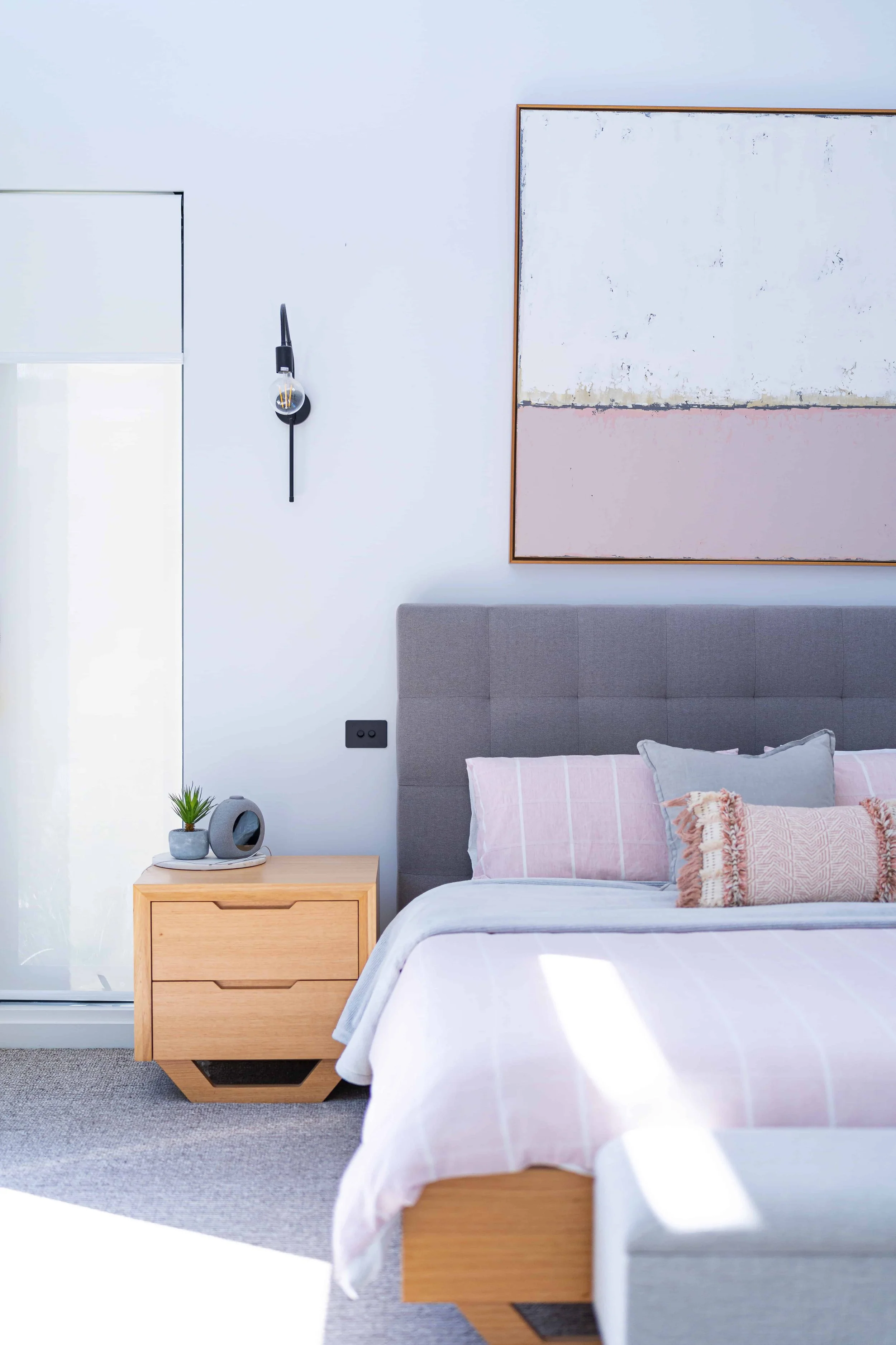 A modern bedroom with a gray upholstered bed, pink and gray pillows, a wooden nightstand with a small plant and a decorative object, a large abstract painting on the wall, and a black wall sconce next to a window with a white roller shade.