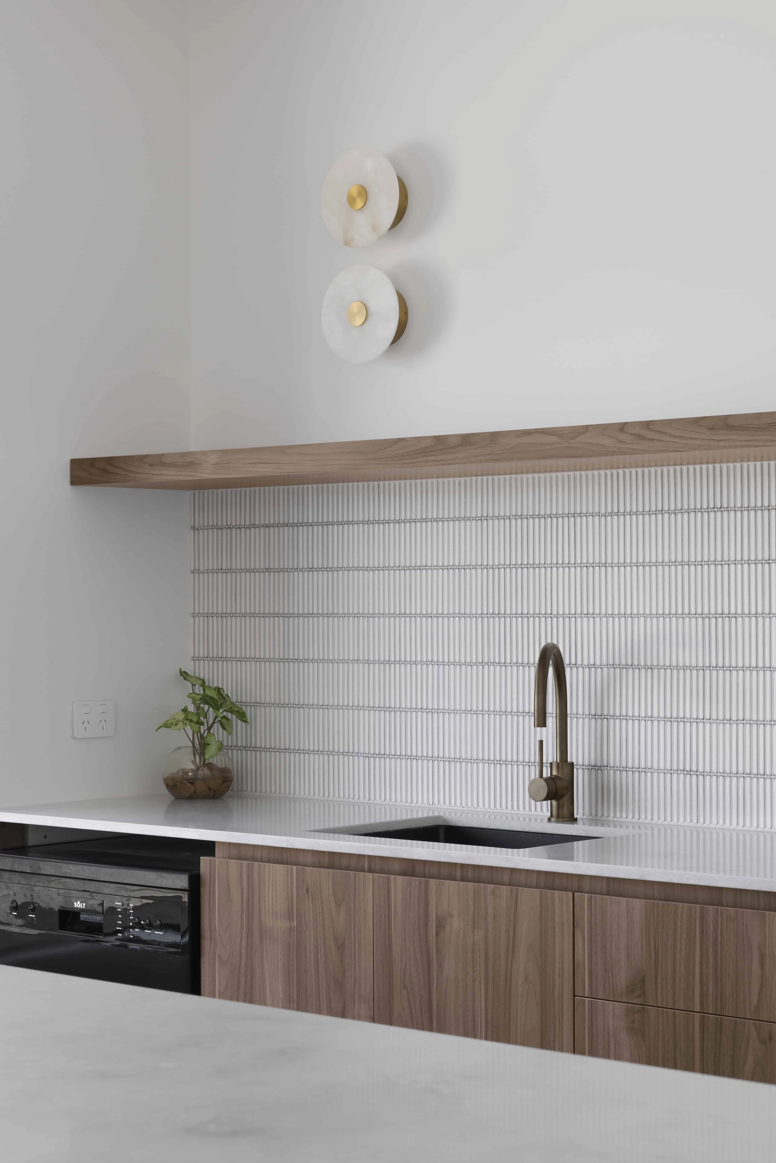 Modern kitchen with white stone benchtop, timber cabinets, and a white finger tile splashback. The wall above has two circular wall sconces with gold accents, and there is a sink with a stainless steel gunmetal faucet.