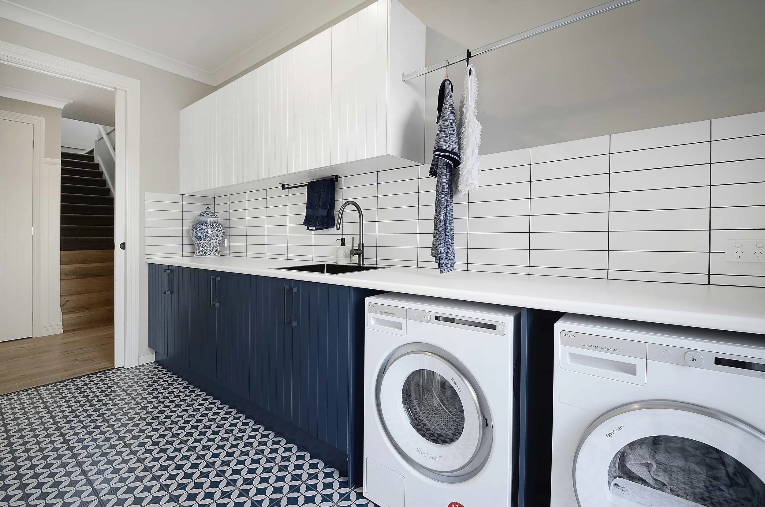 Modern laundry room with white subway tiles, dark blue cabinetry, a laminate benchtop with a rounded edge, and hanging rail.