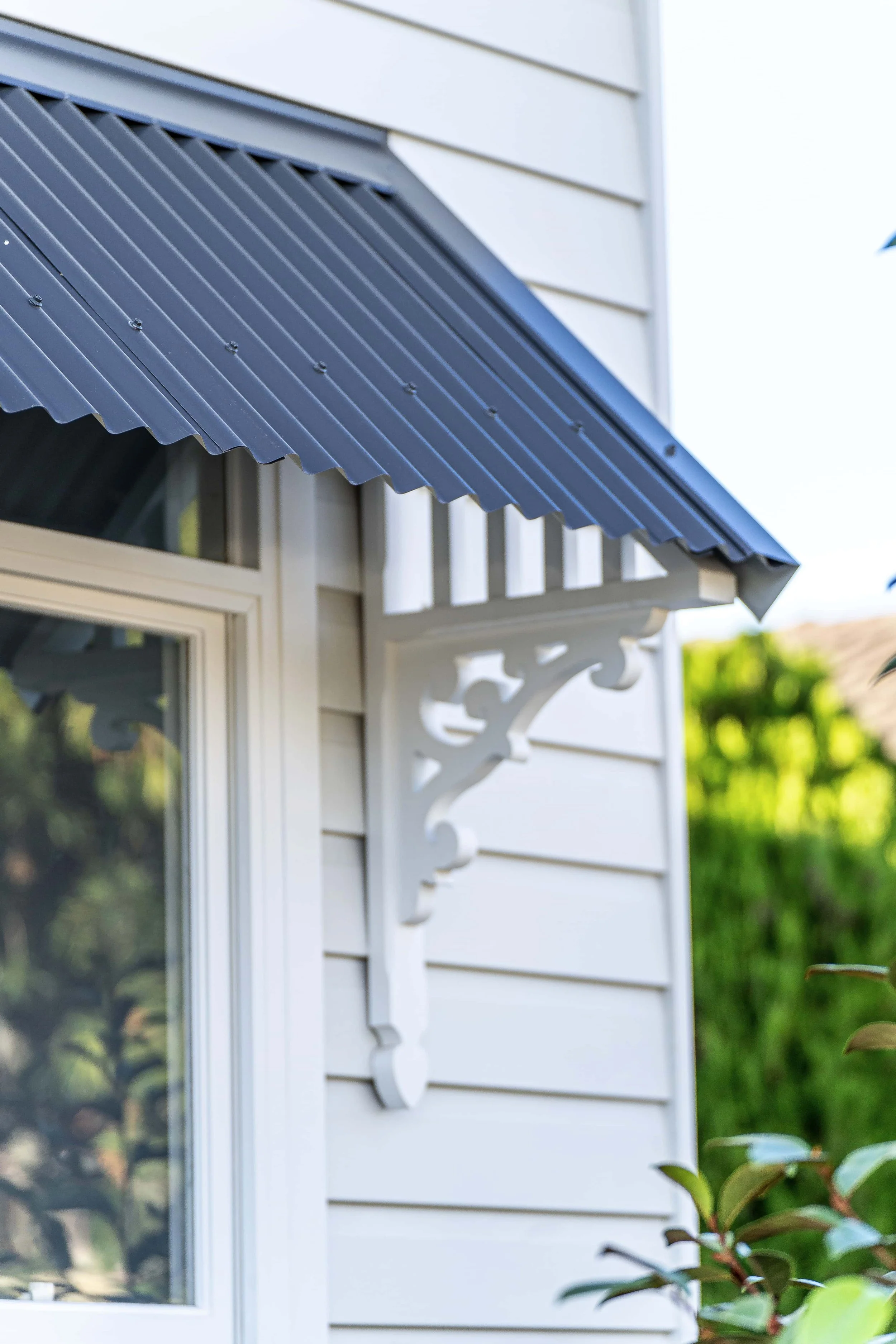 Close-up of a house exterior with a white window, decorative trim, and a dark grey metal awning, surrounded by greenery.
