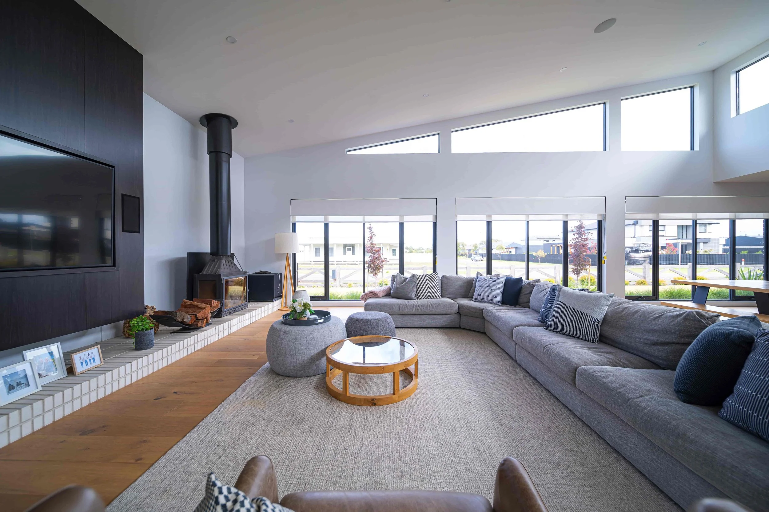 A large open living room with a raked ceiling and clerestory windows. on the left is a feature wall with a wall mounted tv unit in a custom cabinet and a wood log fireplace on a white brick hearth. Natural light floods the space from the windows.