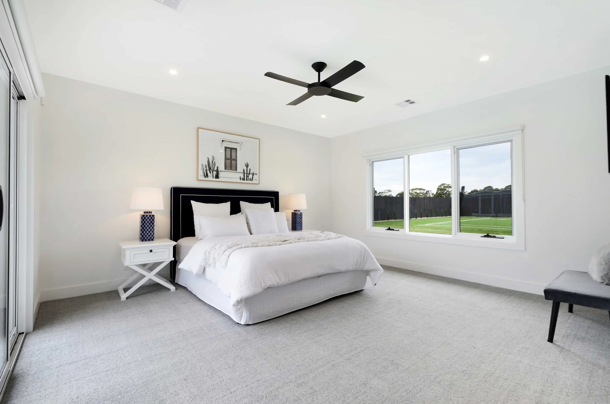 A bedroom with a white bed, two nightstands with lamps, a framed picture above the bed, large windows showing a yard outside, and a ceiling fan.