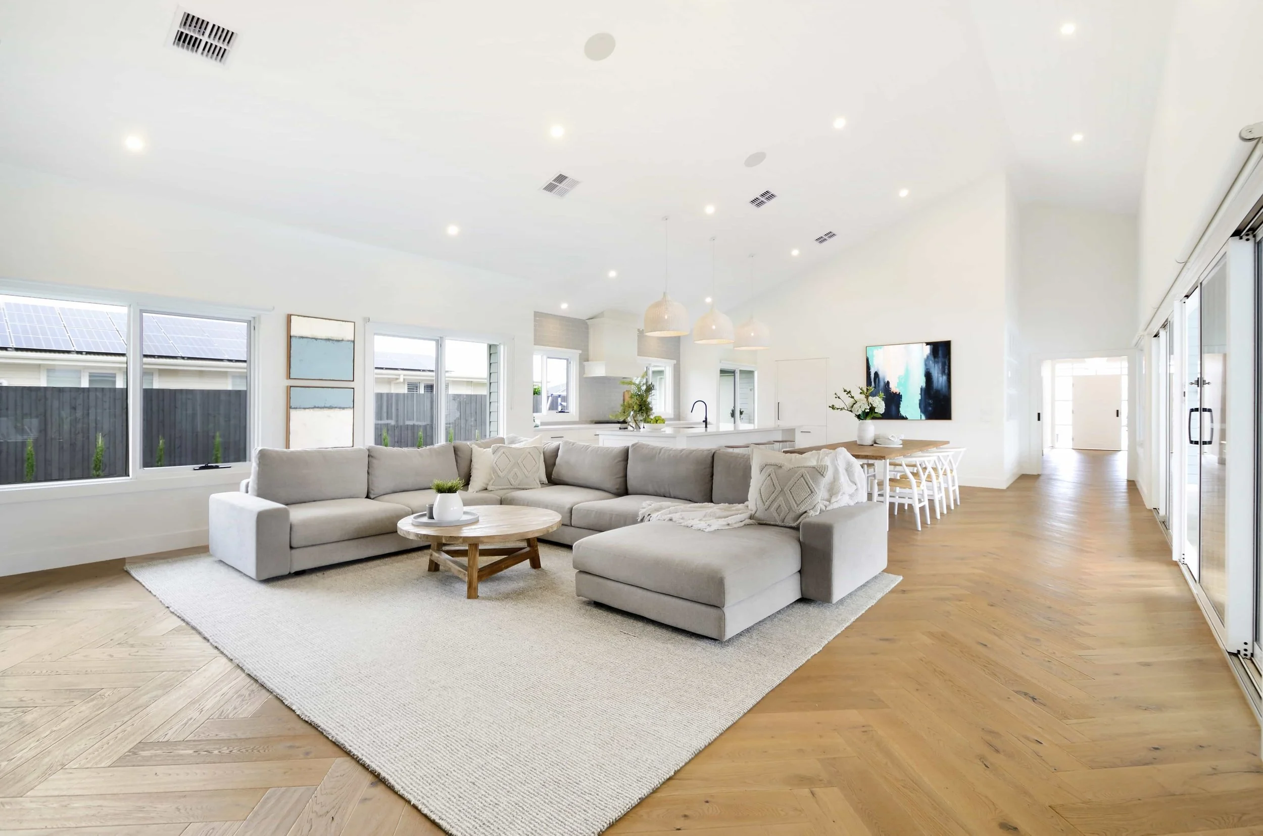 Open concept living room with white walls, large windows, and a view into the kitchen with white cabinets and plants. There is a cathedral ceiling with an off centre ridge line.