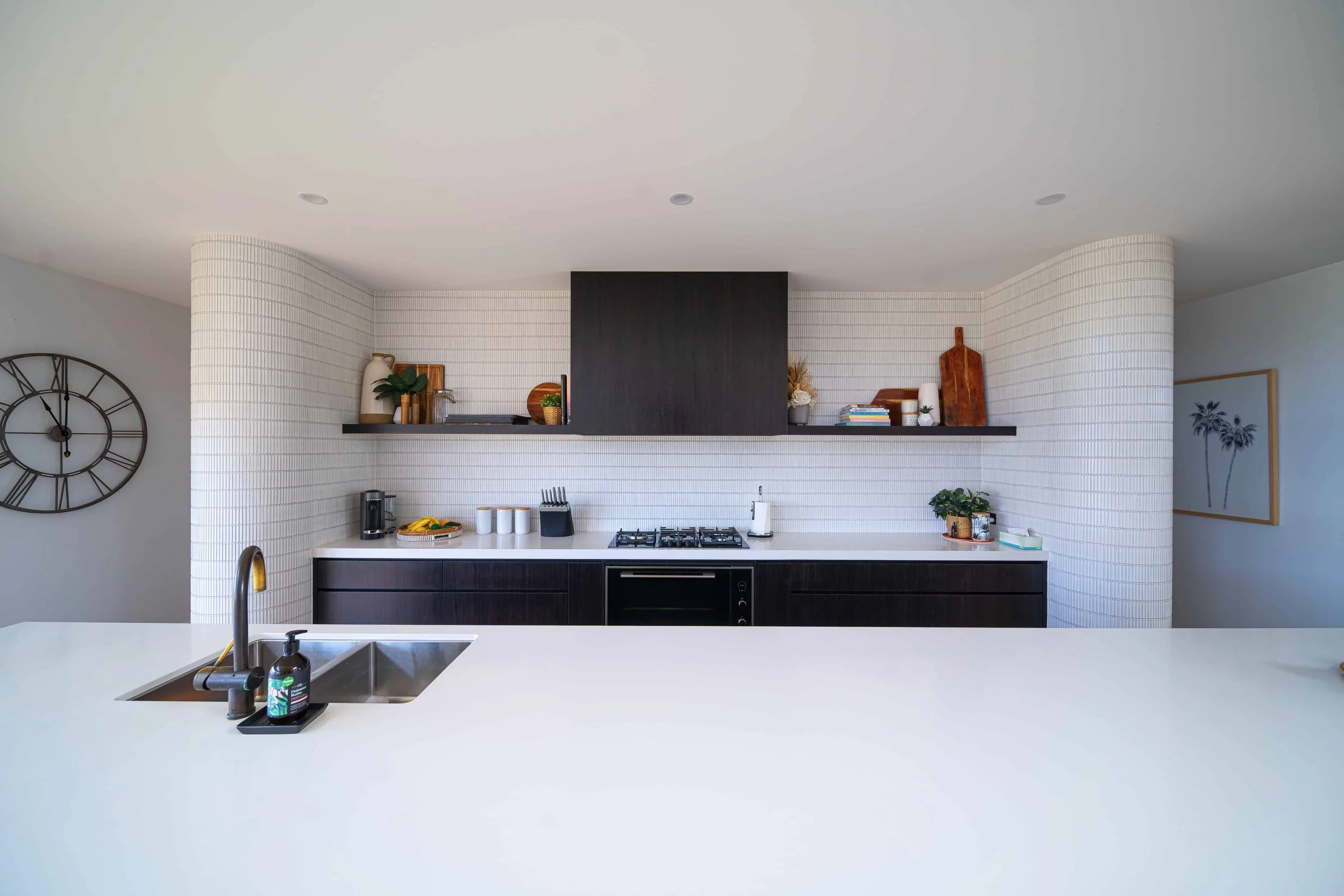 Modern kitchen and island with a white benchtop, black cabinets, and a white finger tile splashback. There are curved feature piers on either side of the back kitchen bench