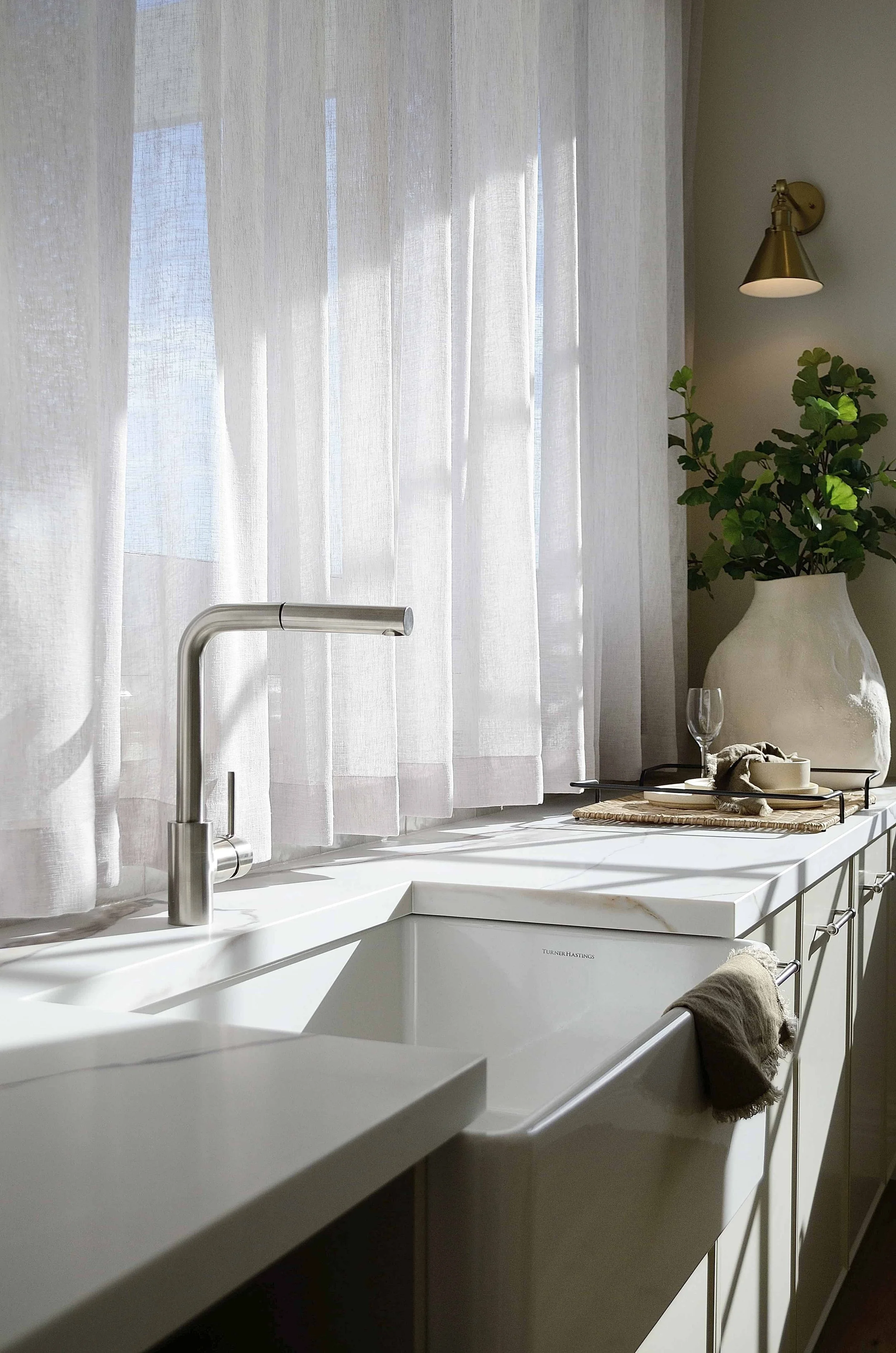 Butler's kitchen sink with white curtains, silver faucet, a large white vase with greenery, a tray with a wine glass and bowl, and a towel on the countertop, in natural daylight.