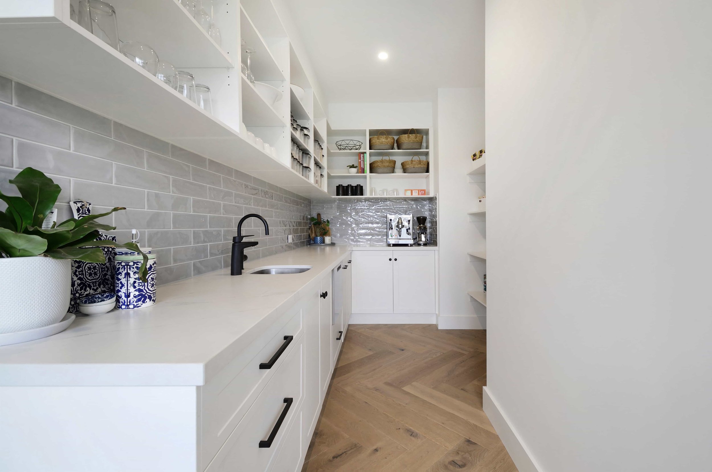 White butler's pantry with open shelves overhead, glossy grey subway tile splashback, black cabinet drawer handles, black faucet, and timber herringbone flooring.