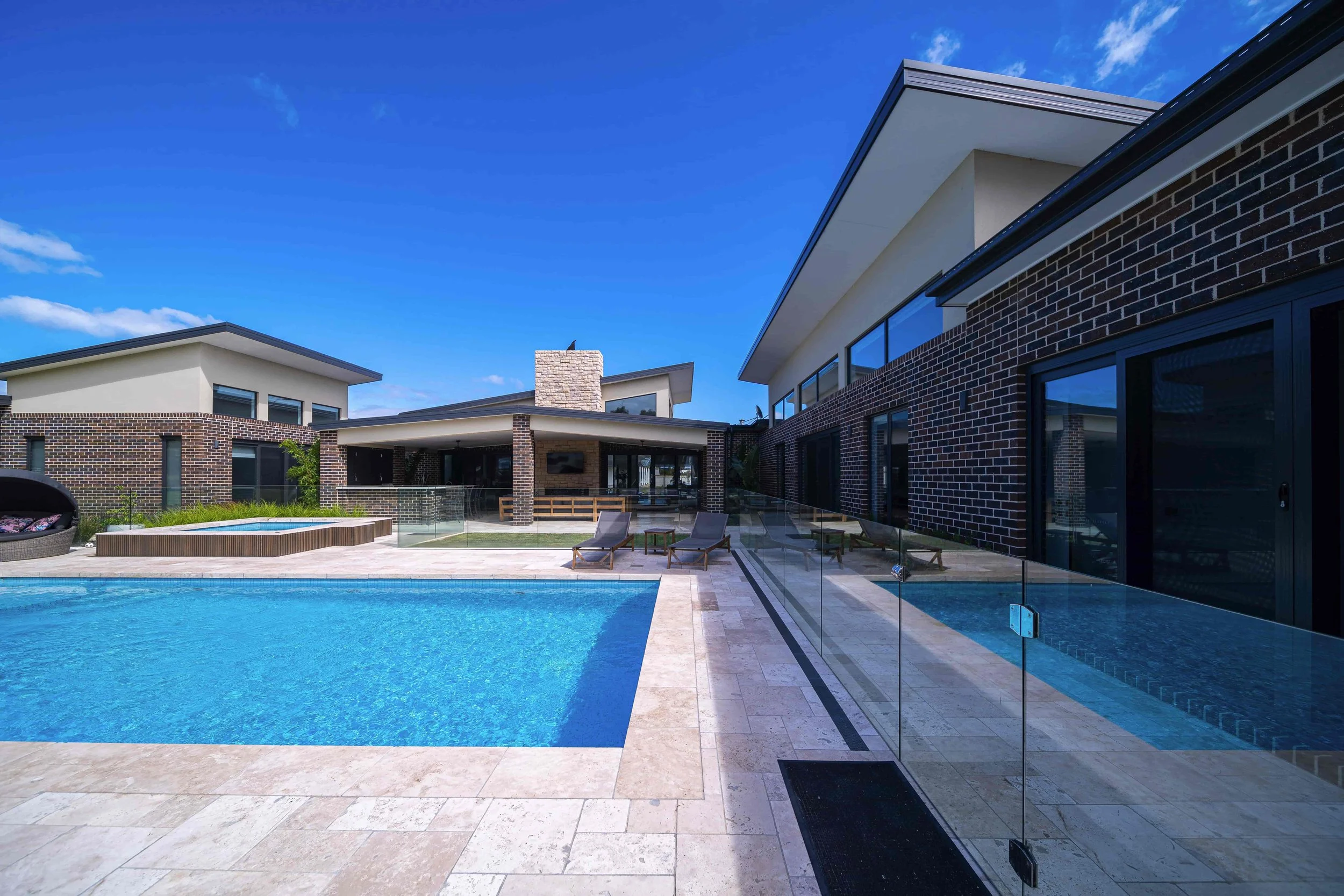 Modern house with brick and rendered white walls, large windows, and skillion roofs. A swimming pool with a glass barrier surrounds a central paved outdoor area with lounge chairs and outdoor furniture.