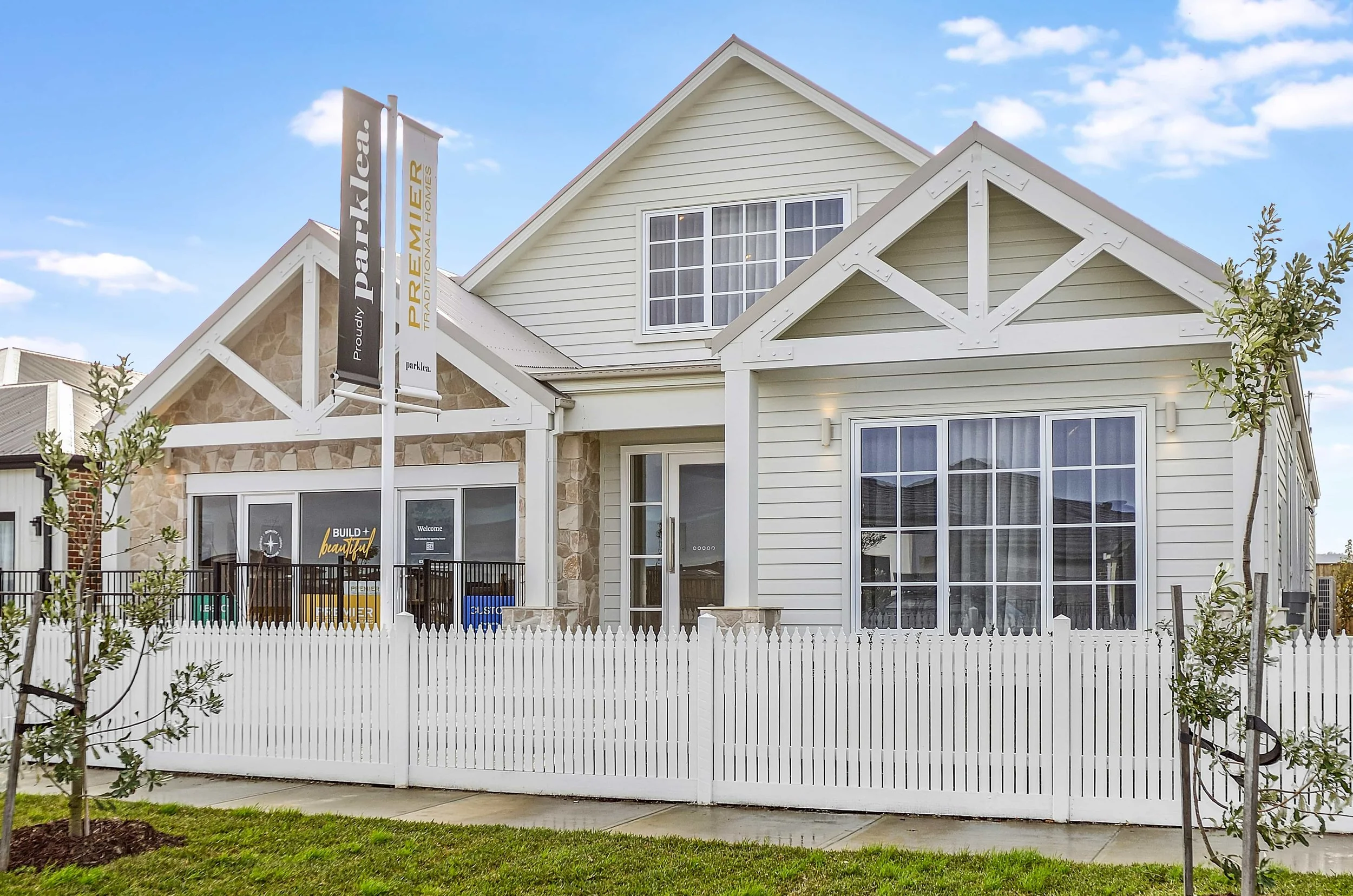 New house with white weatherboard exterior, stone accents, large colonial front windows, and a white picket fence, under a blue sky with scattered clouds.