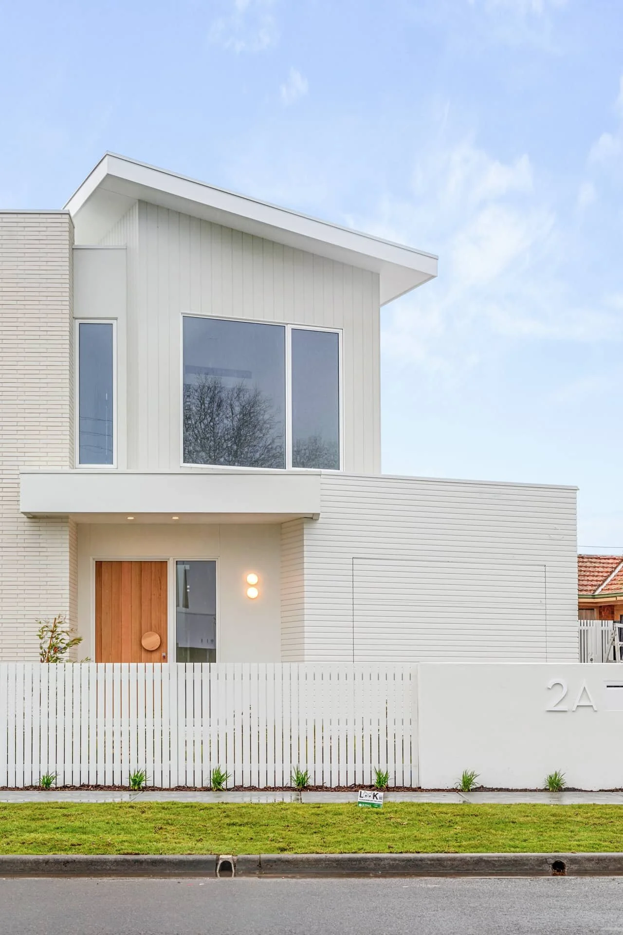 Contemporary two-storey residential home with a white brick and weatherboard exterior, large offset windows, timber entry door, white picket fence, and well-maintained lawn.