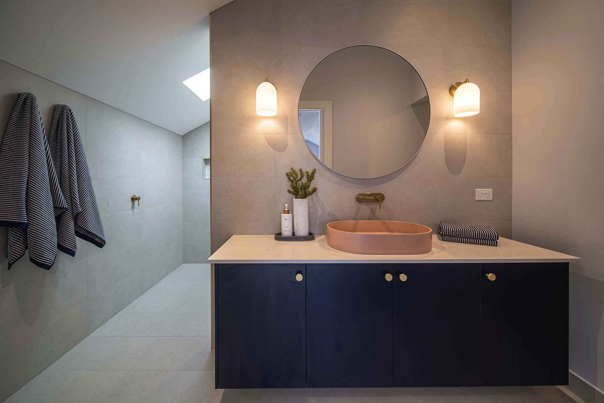 Modern bathroom with a navy blue vanity featuring a round, pink above bench basin, a large round mirror, two wall-mounted light fixtures either side, with a neutral colour scheme and minimalist decor.