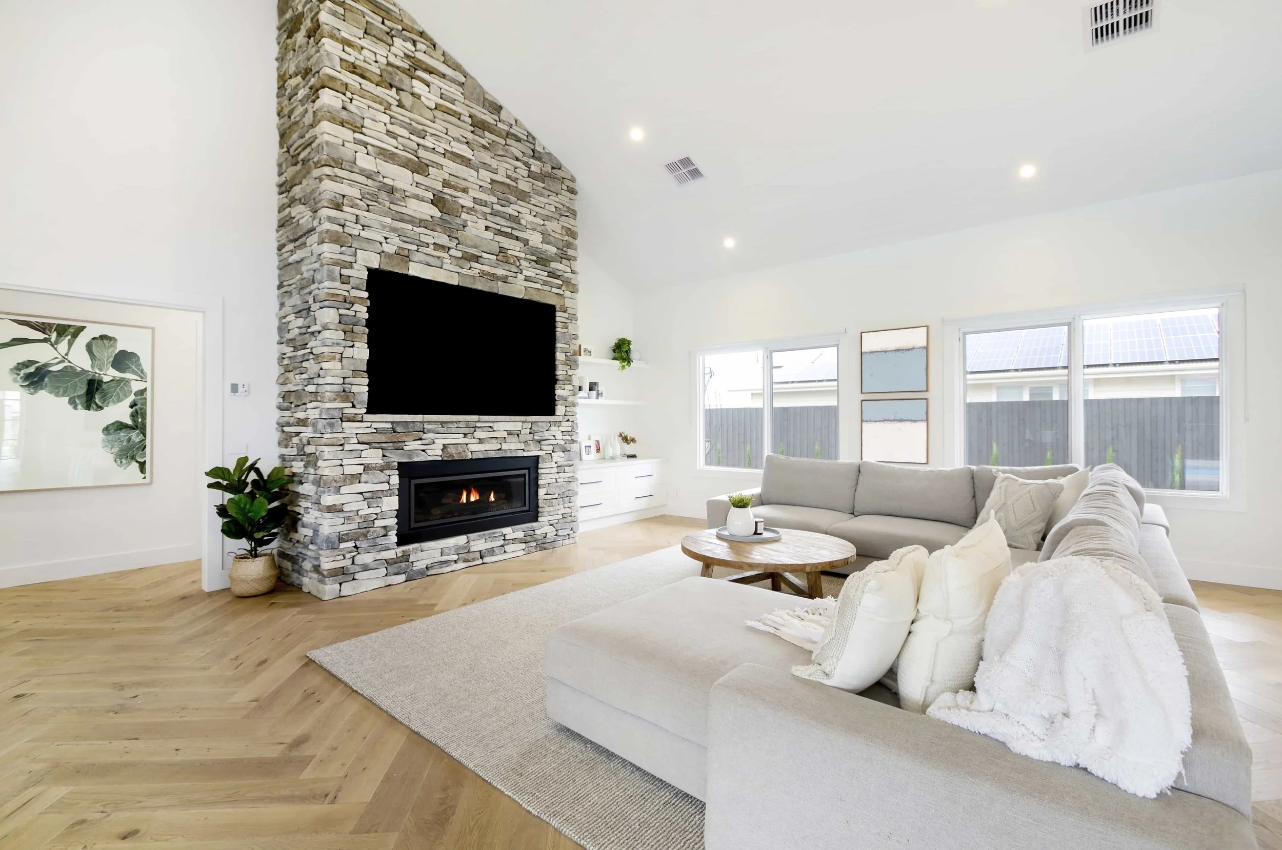 Living room with a stone clad fireplace, beige sofa, wooden coffee table, large windows, and decorative plants.