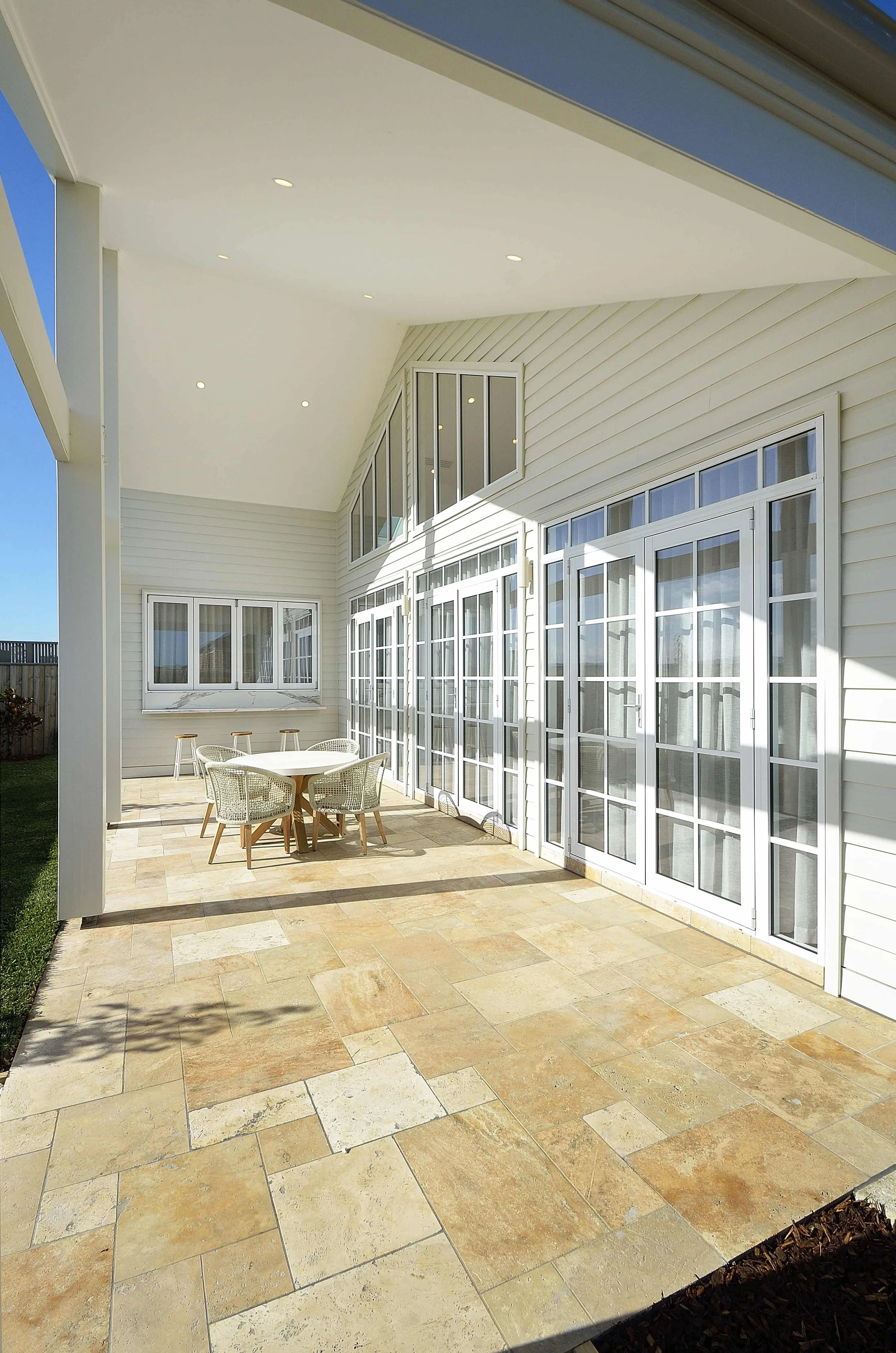 An alfresco area with a table and four wicker chairs on tan stone tiles, adjacent to a house with large colonial glass doors and windows, white weatherboards, and a white cathedral ceiling with small recessed downlights.