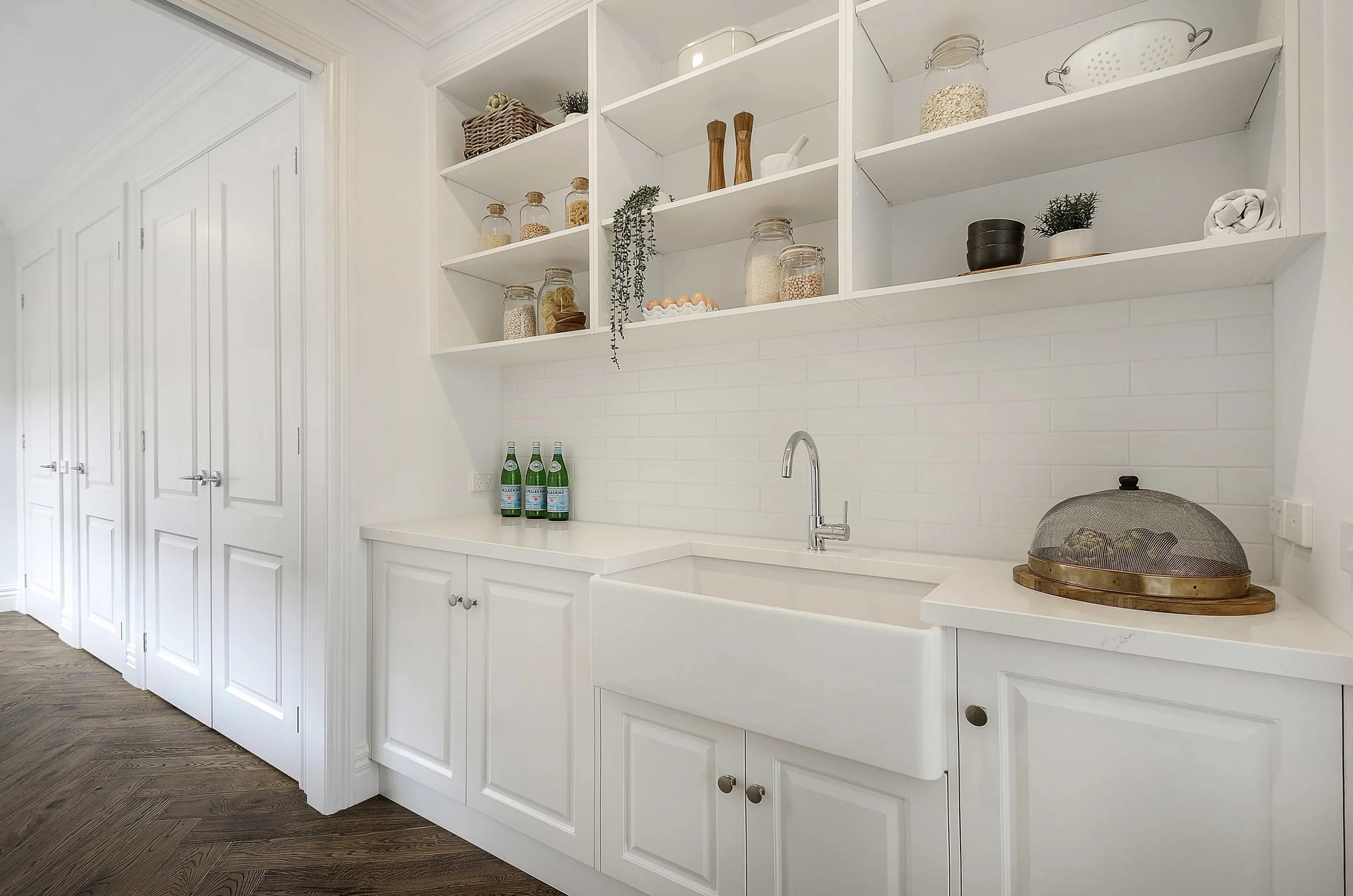 A modern butler's pantry with white routed door panels and open shelving on the wall above. The white stone bench features a white farmhouse sink, chrome faucet, and white horizontal subway tiles.