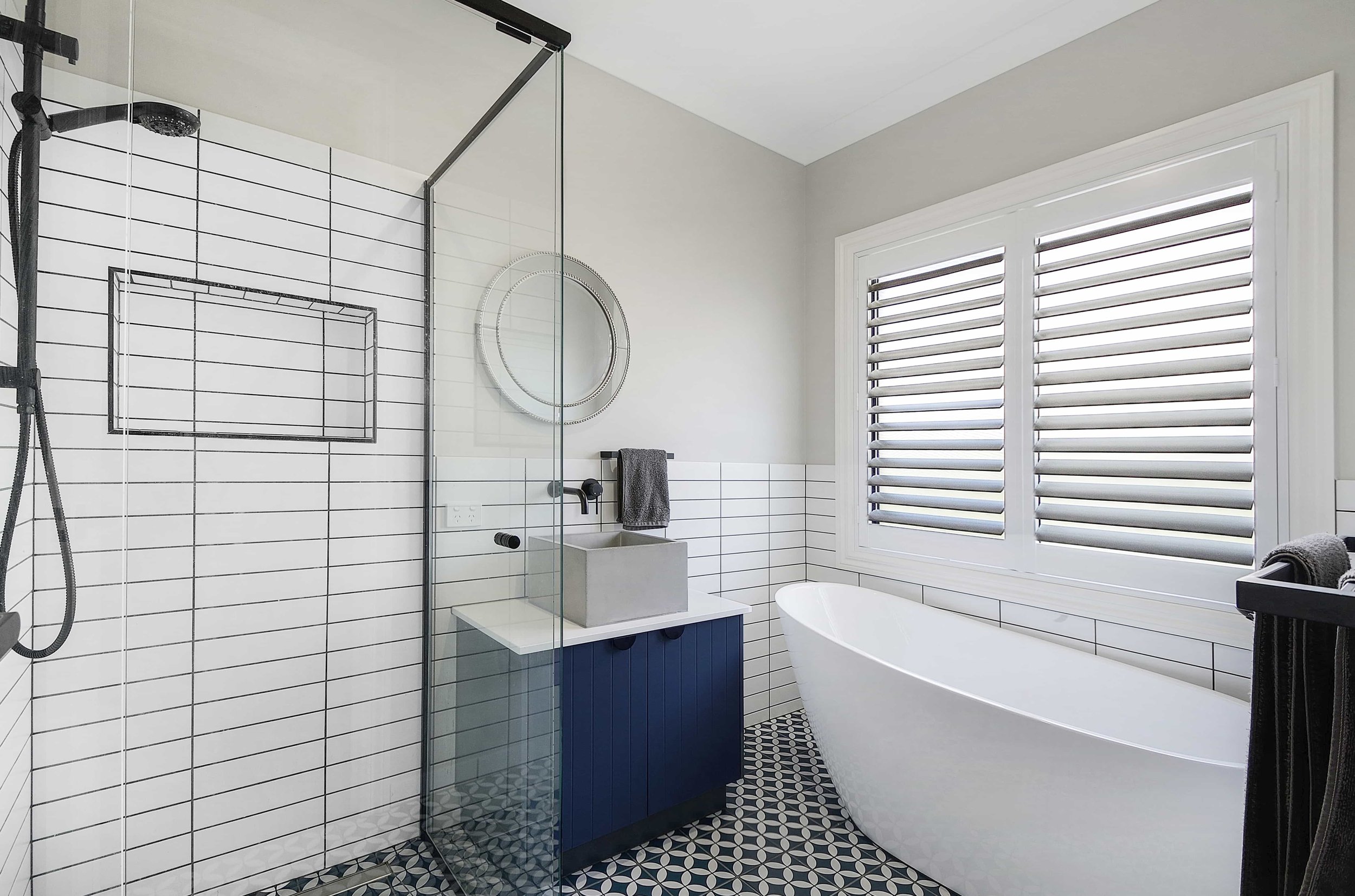 Modern bathroom with white subway tiled walls, a navy and white patterned floor, a glass-enclosed shower area, a white freestanding bathtub, a blue vanity with a concrete sink, a round mirror, and large window with white horizontal blinds.