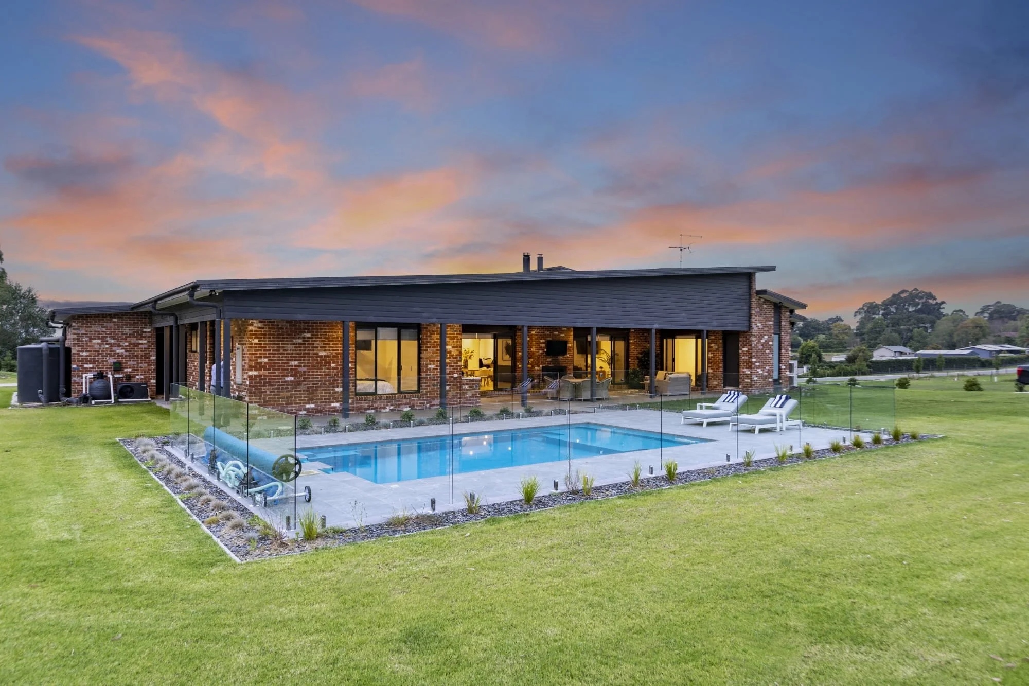 Modern red brick house with large glass doors and windows overlooking a backyard with a swimming pool, lounge chairs, and a well-maintained lawn, under a colourful sunset sky.