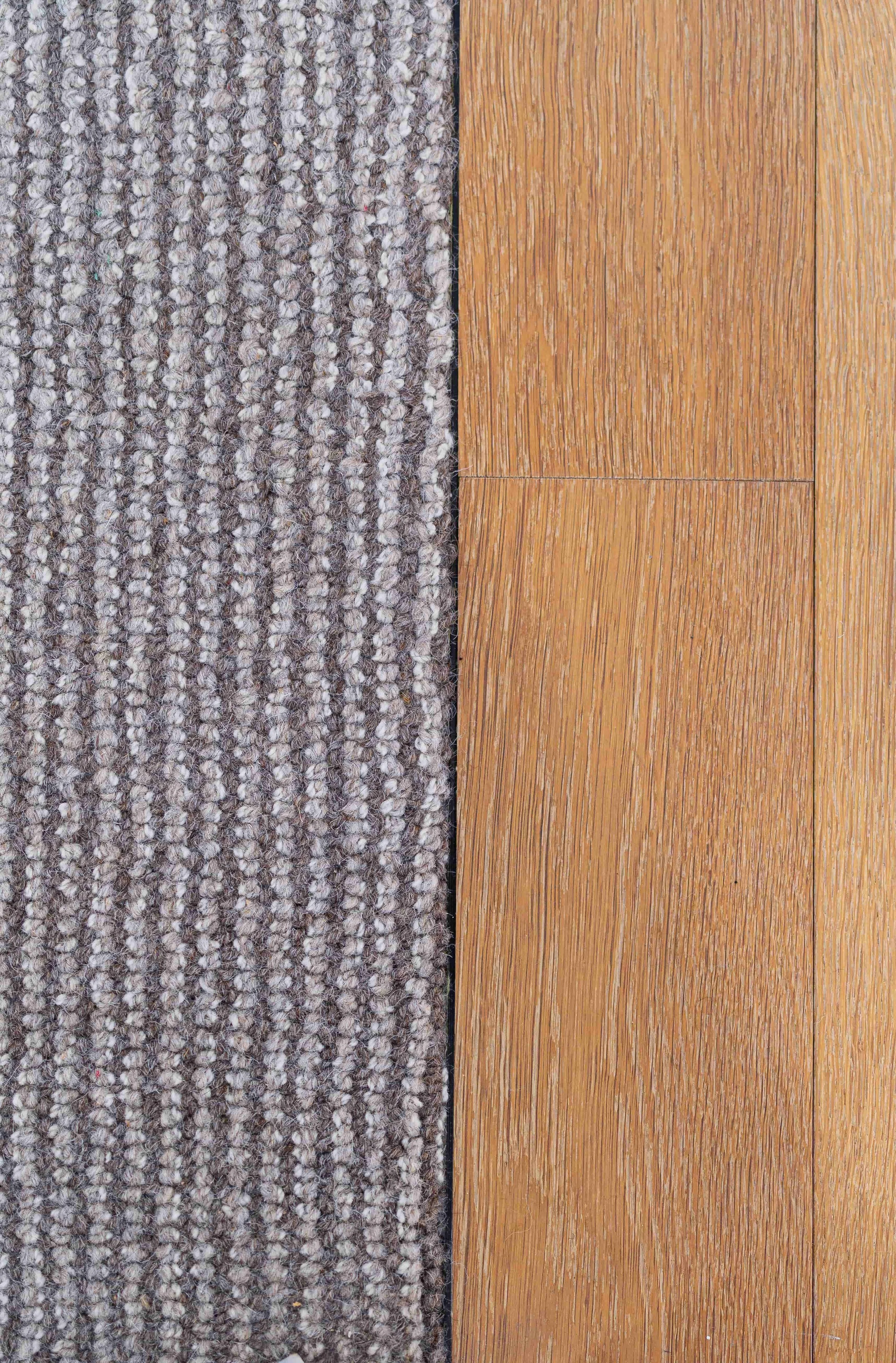 Close-up of a textured gray carpet on the left side and a hardwood floor with a reddish-brown tone on the right side.