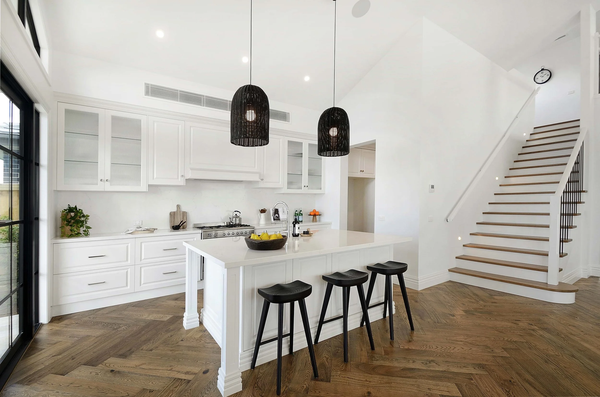 Modern kitchen with routed door panelling, an island bench with traditional detailing, and a staircase ascending to the right. The floor is in a dark timber herringbone pattern.