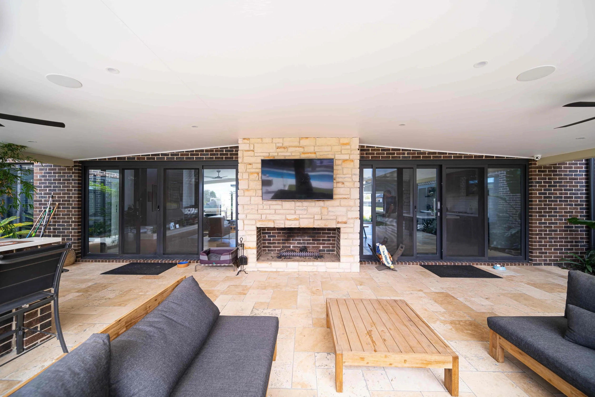 An outdoor alfresco area with a feature stone fireplace. The cream coloured floor tiles are in a random pattern. Large black framed sliding doors lead to the interior.
