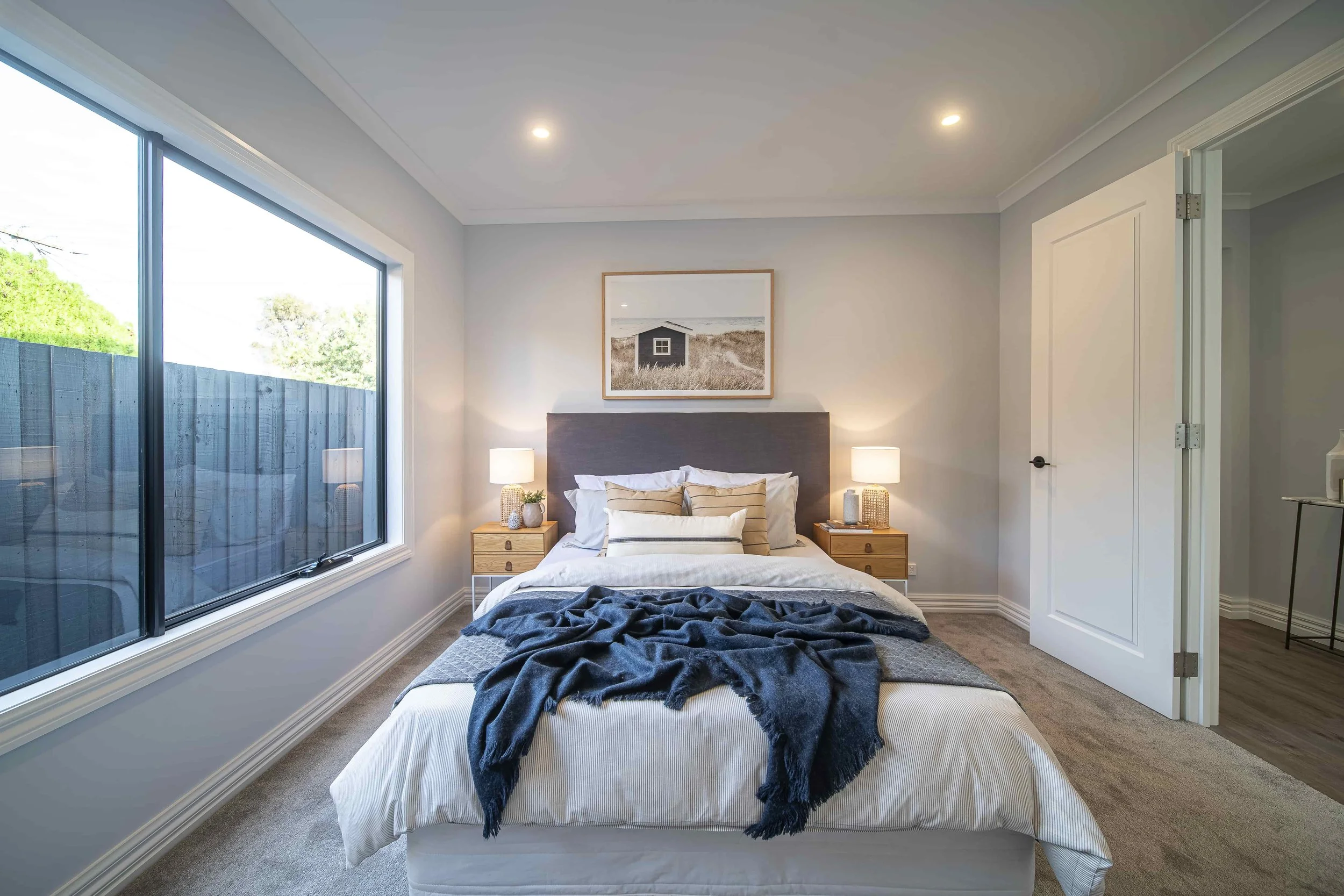 A modern bedroom with a large window to the left, featuring a bed with a white bedspread, striped pillows, and a dark blanket. There are two matching wooden nightstands with lamps and decorative items. A framed black and white photo of a house on a b