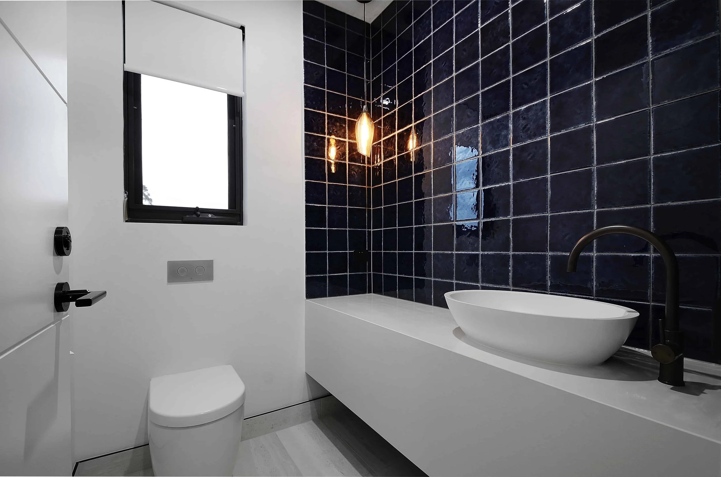 A powder room with navy blue tiled wall, white floating stone vanity with an oval above bench basin, black faucet, concealed toilet, window with white blind, and a hanging pendant light.