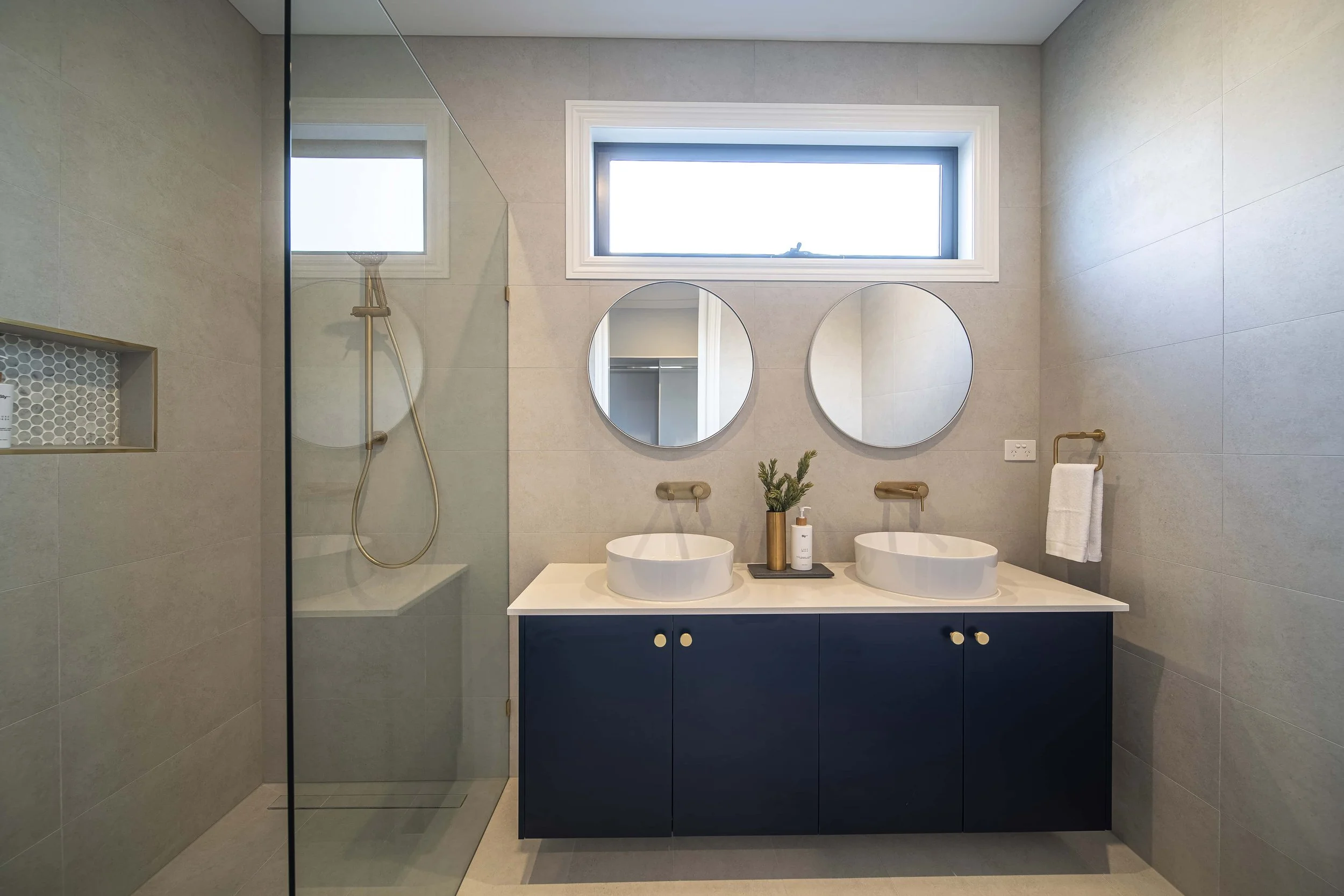 Modern ensuite with two round basins, round mirrors, small plant, soap dispenser, and towel rack. The walk in shower is partially enclosed with glass. The wall mounted shower rail, vanity handles, and tapware are gold plated.
