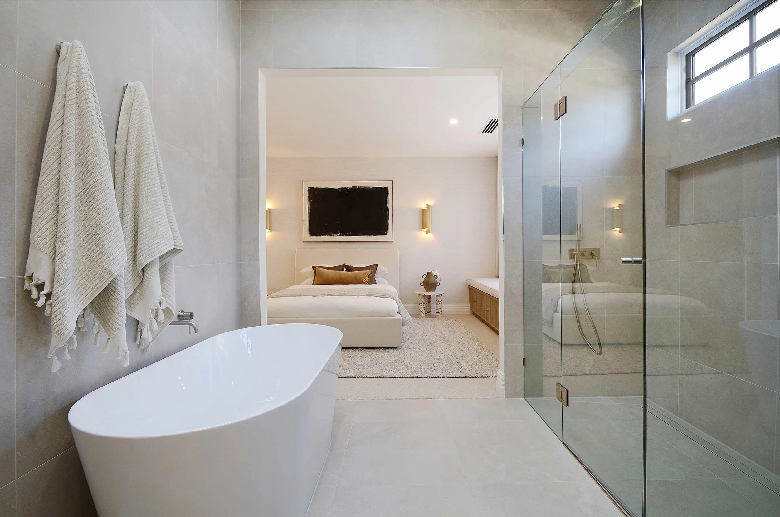 Modern ensuite with a freestanding bathtub, two white towels hanging on the wall, a glass shower enclosure, and a bedroom visible through an open doorway. The bedroom has a bed with pillows, a painting, and feature bedside wall lamps.
