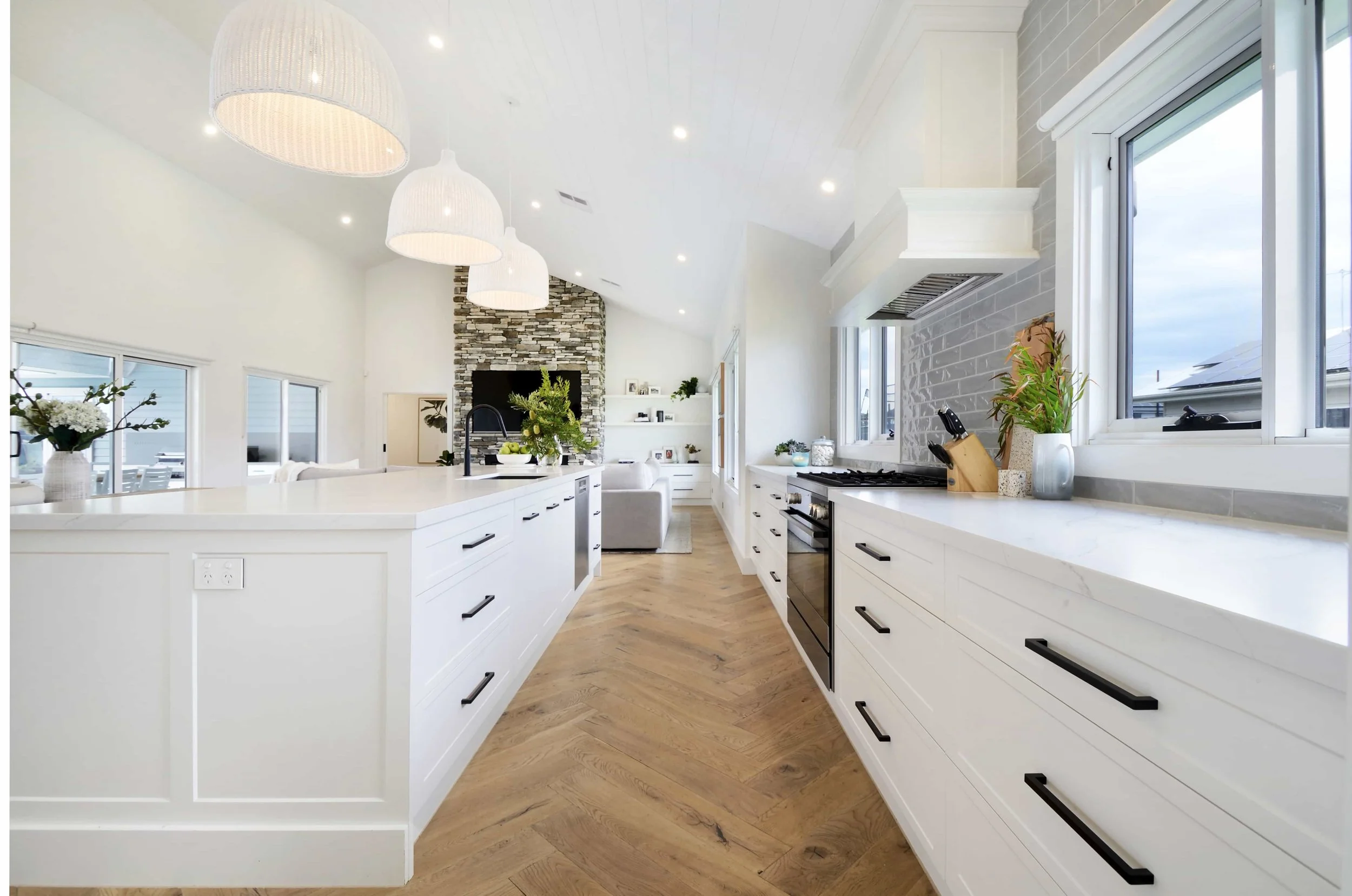 Modern white kitchen with black hardware, large windows, and a stone fireplace in the adjacent living area.