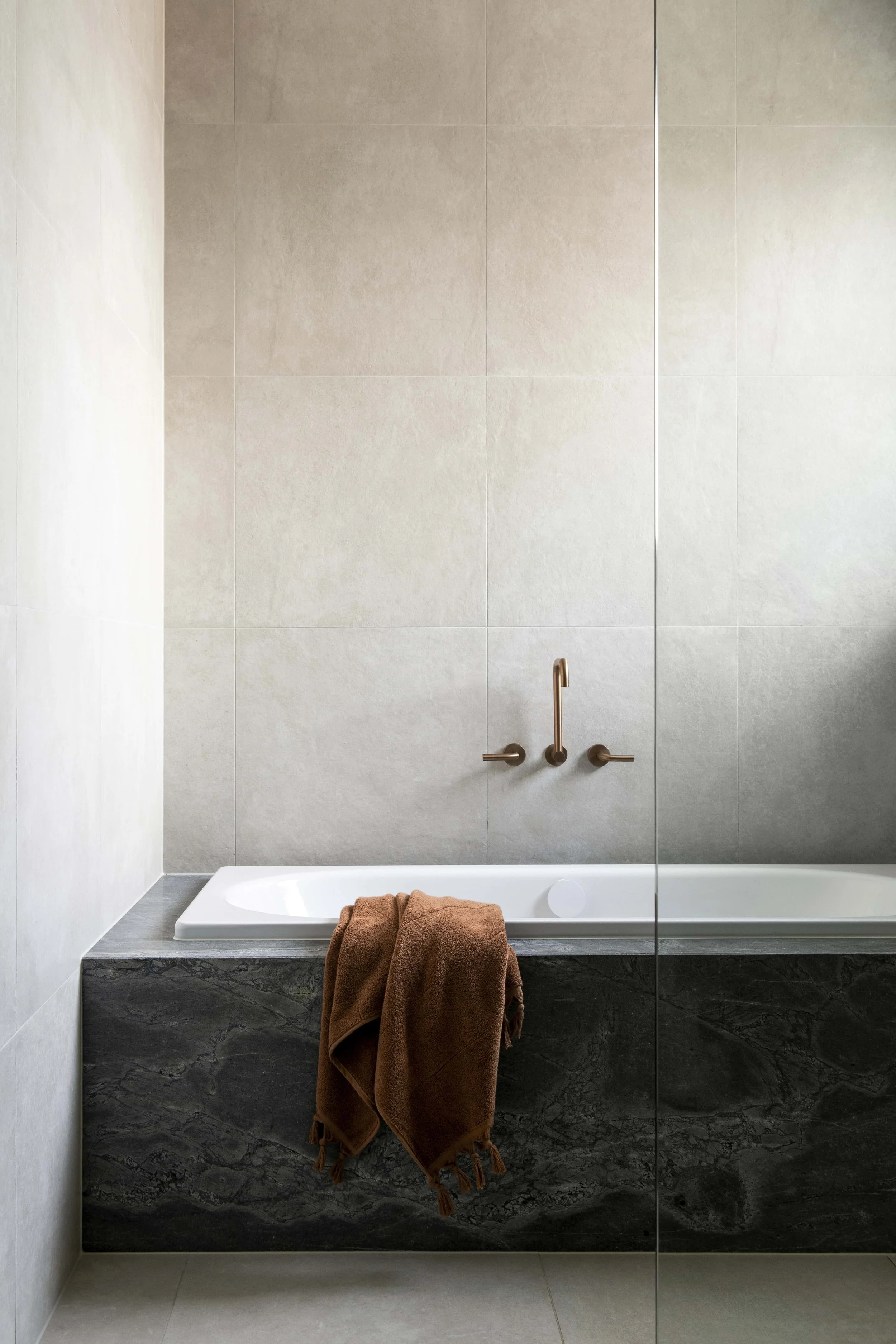 Modern bathroom with beige tiled wall, white bathtub, dark grey stone hob, bronze tapware, and brown towel hanging over the edge of the tub.