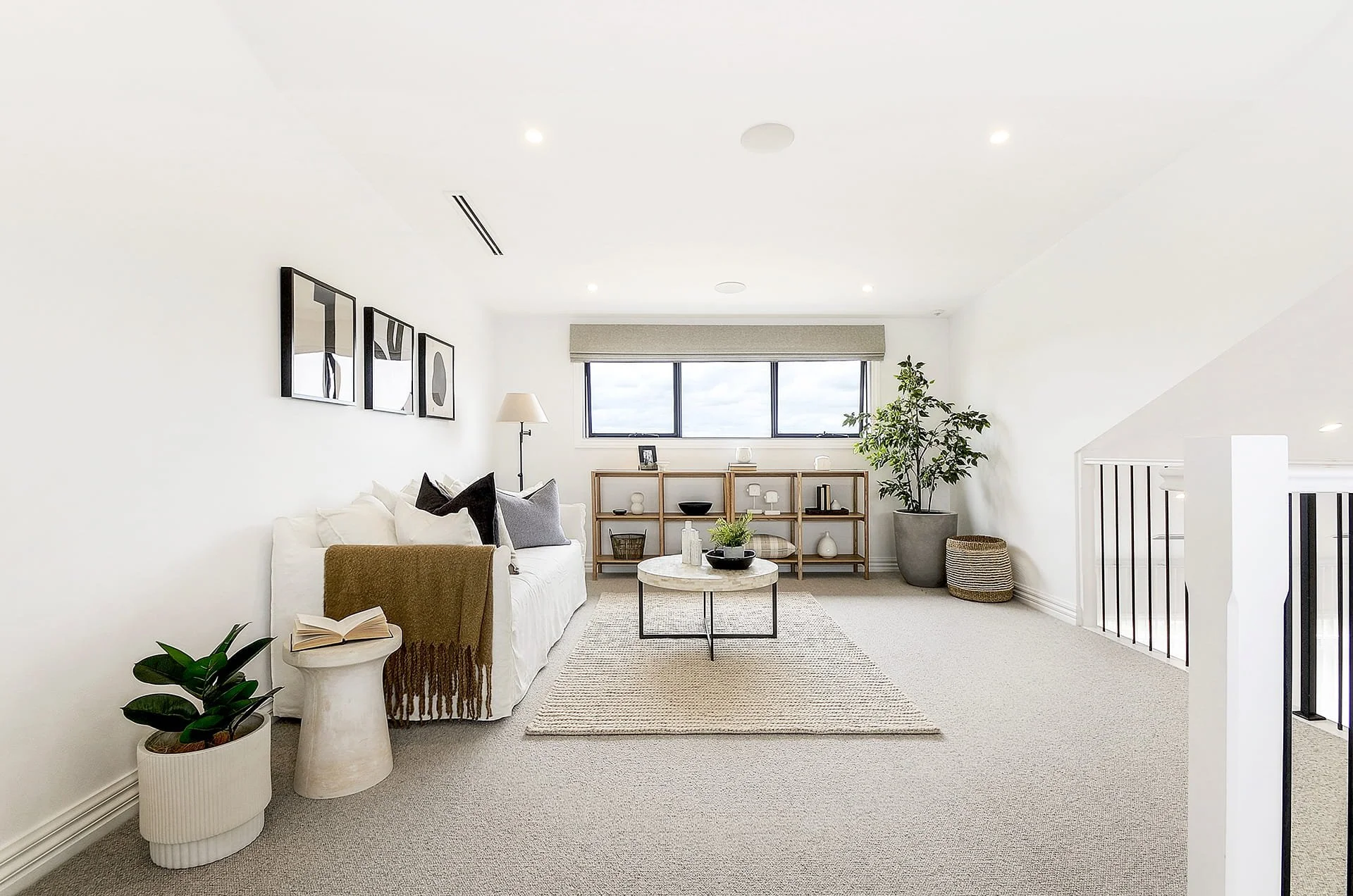 Bright, modern living room with white walls, large window, and minimalist decor including a white sofa with gray and black pillows, a wooden bookshelf, a potted plant, and framed abstract art.