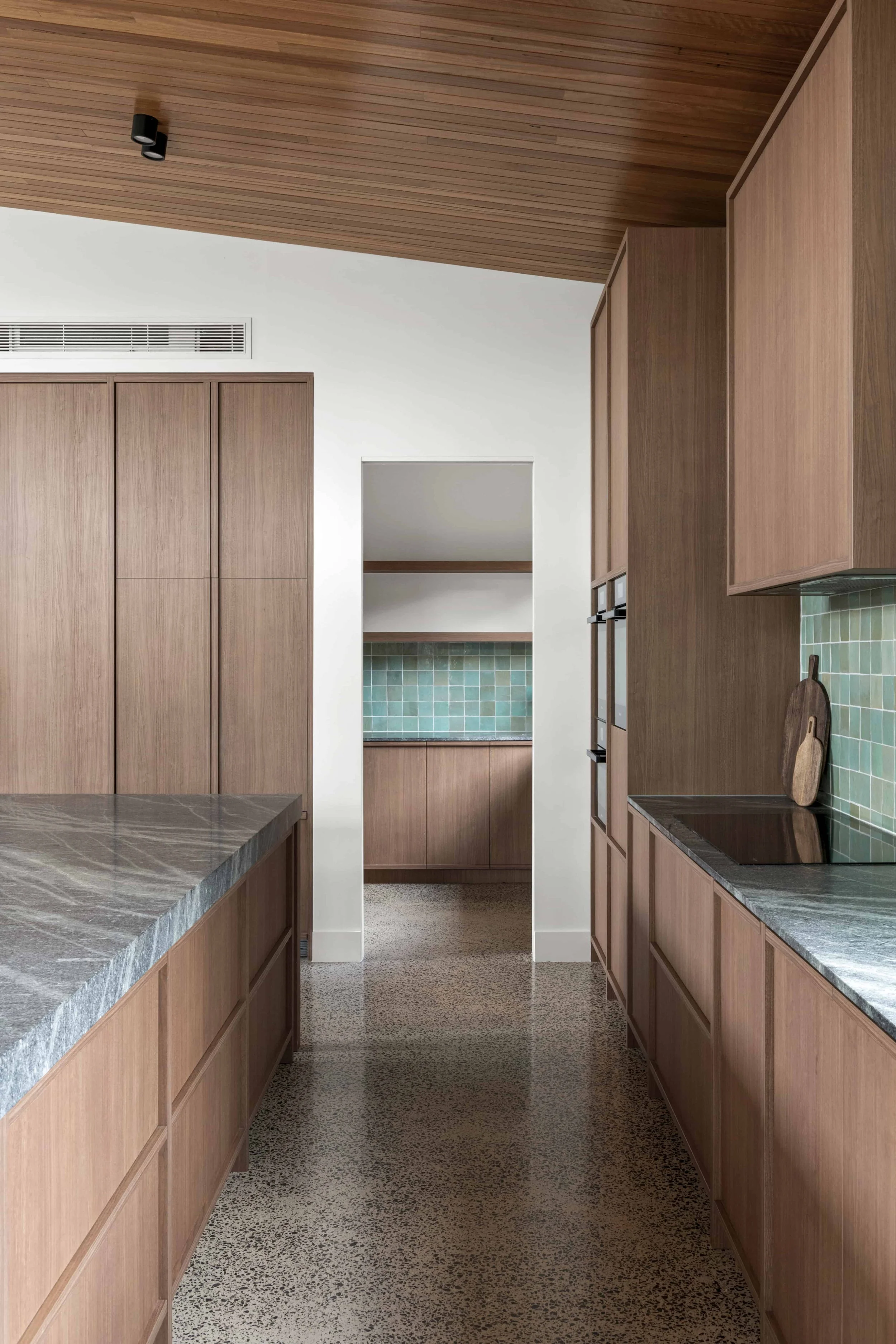 Interior view of a modern kitchen with timber cabinets, grey stone benchtops, green mosaic tile splashback, and a polished concrete floor, shown from the perspective of a narrow hallway.