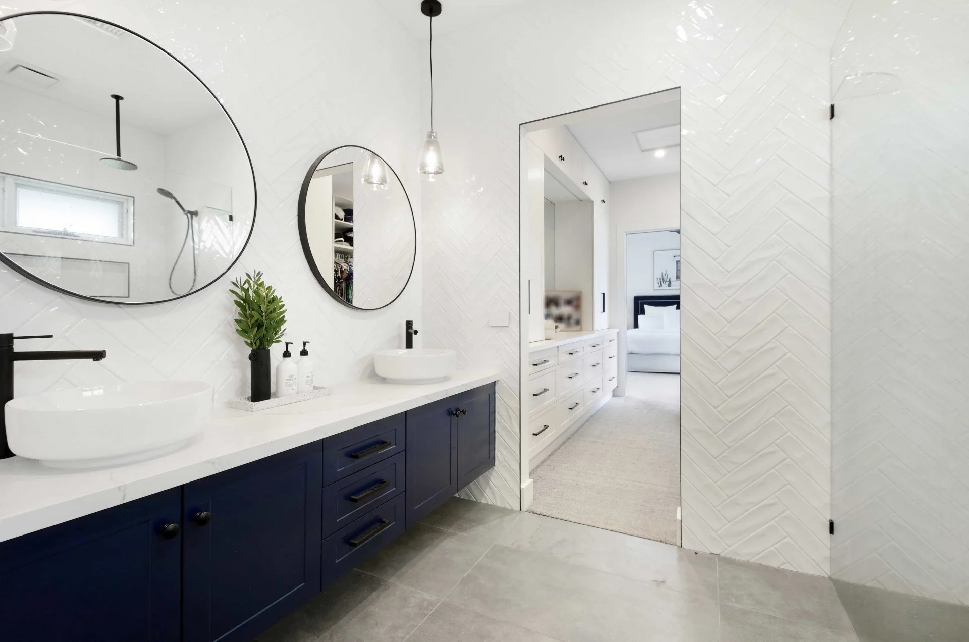 Modern ensuite with a navy blue vanity, white stone benchtop, and two round above counter basins. Glossy subway tiles are laid in a herringbone pattern on the wall going up to the ceiling. The room looks out to the adjoining walk in robe.