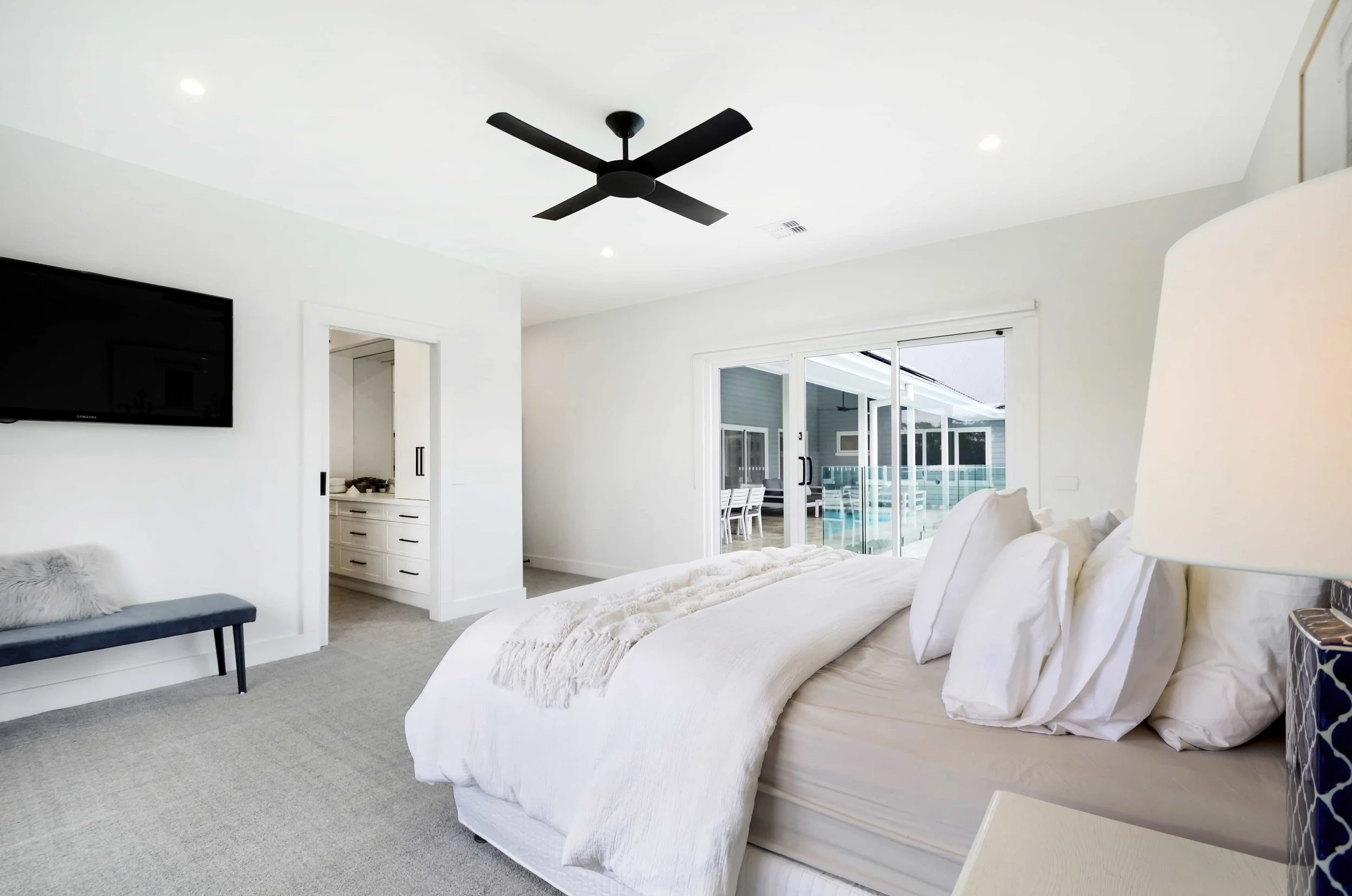 A master bedroom suite with sliding glass doors leading to an outdoor patio with pool, and a mounted TV on the wall. A cavity door across the bed opens into the adjoining walk in robe.