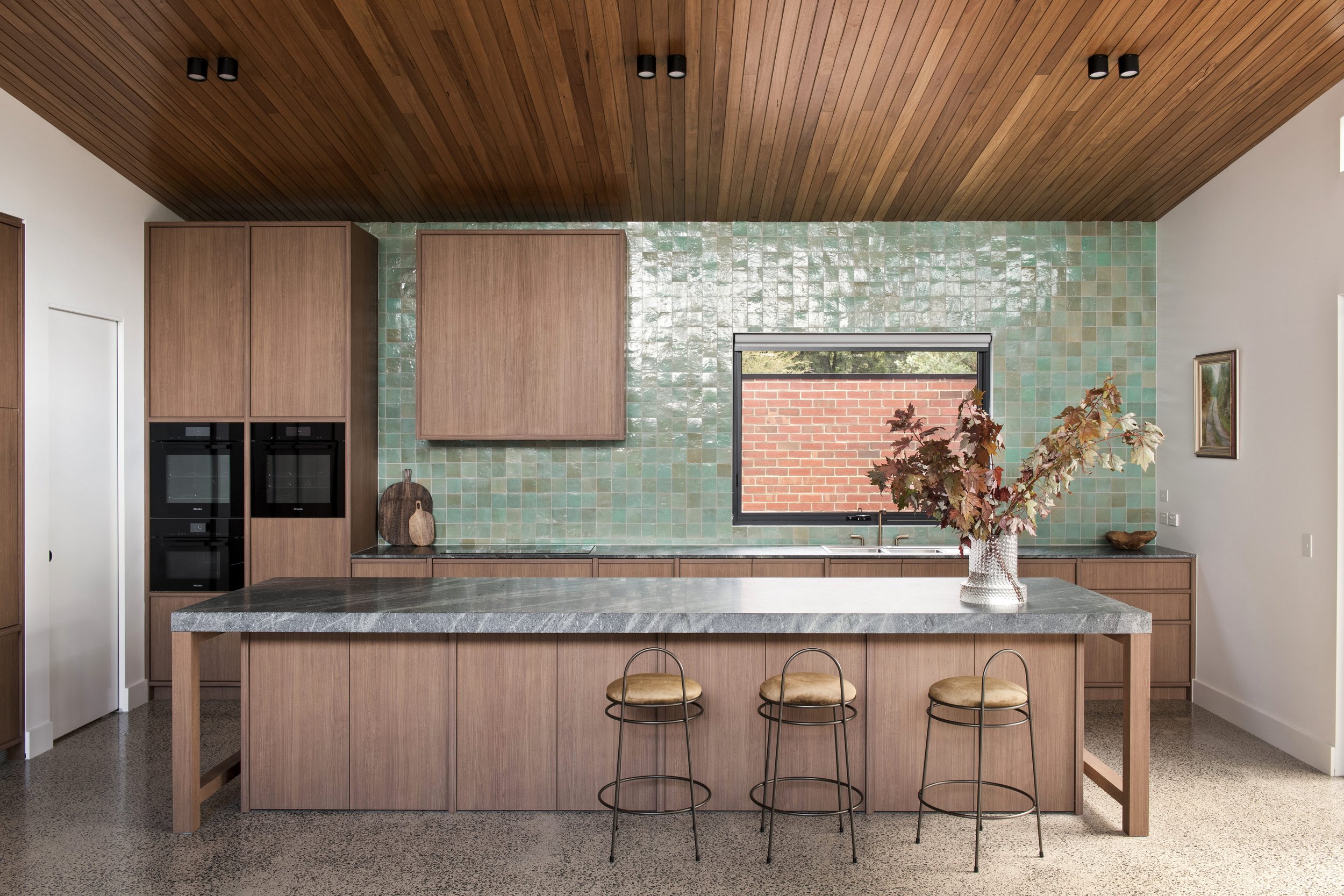 Modern kitchen with wood cabinets, green backsplash tiles, a window with a brick wall outside, a kitchen island with bar stools, and a decorative vase with leaves.