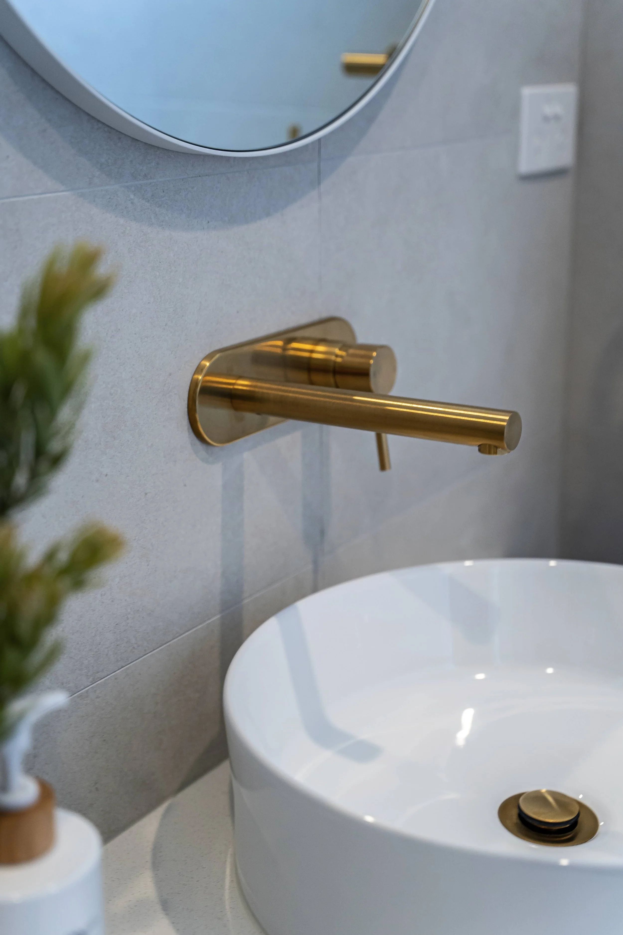A close up of a modern bathroom vanity with a round above bench basin, a gold faucet and basin drain, and a small vase with a plant. There is a round mirror and a wall outlet on a grey tiled wall.