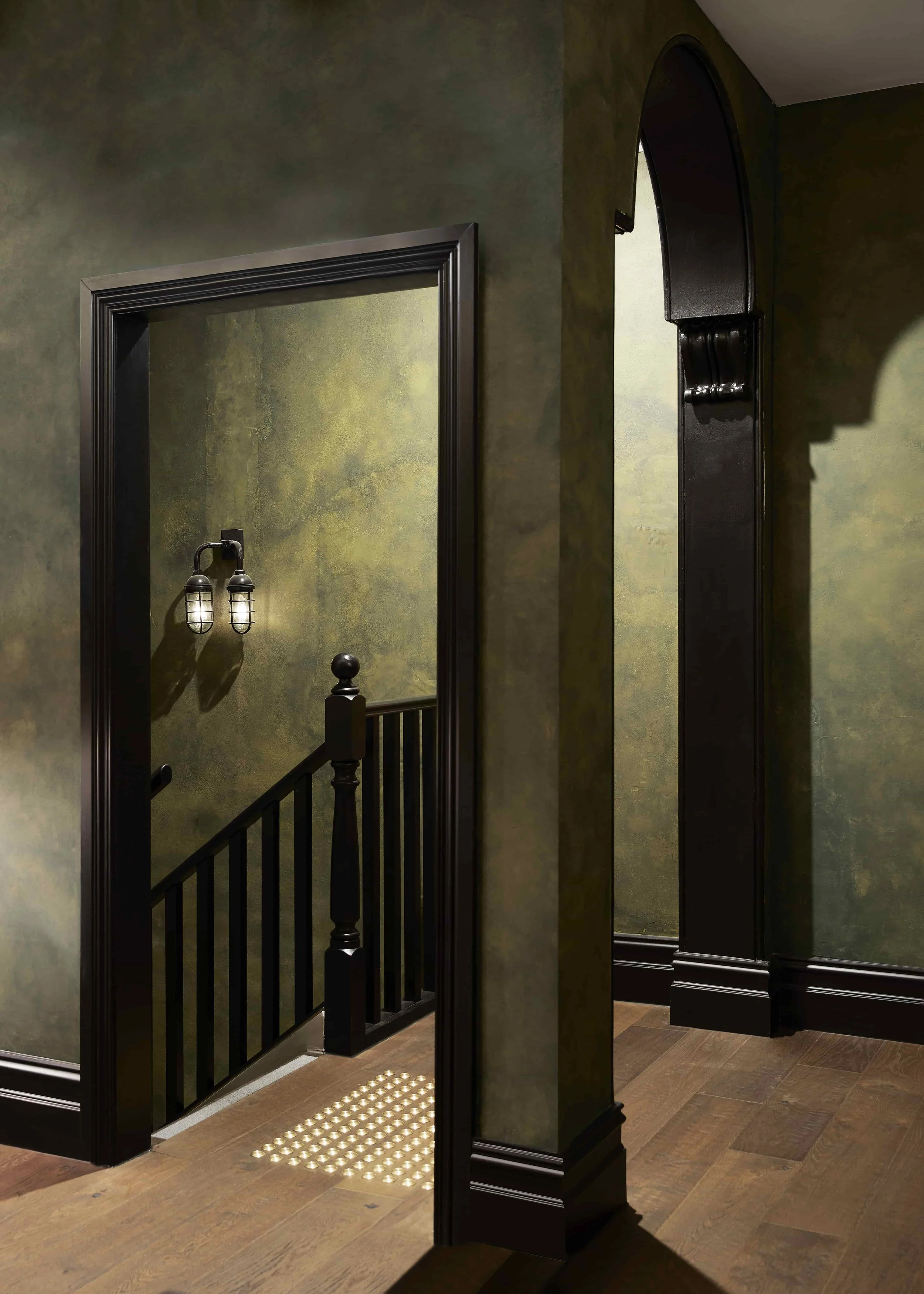 Interior view of a staircase with black railing next to a dappled dark green wall, and decorative vintage-style lighting fixtures. The floors are a rustic timber with a tactile paving mat at the staircase entrance.
