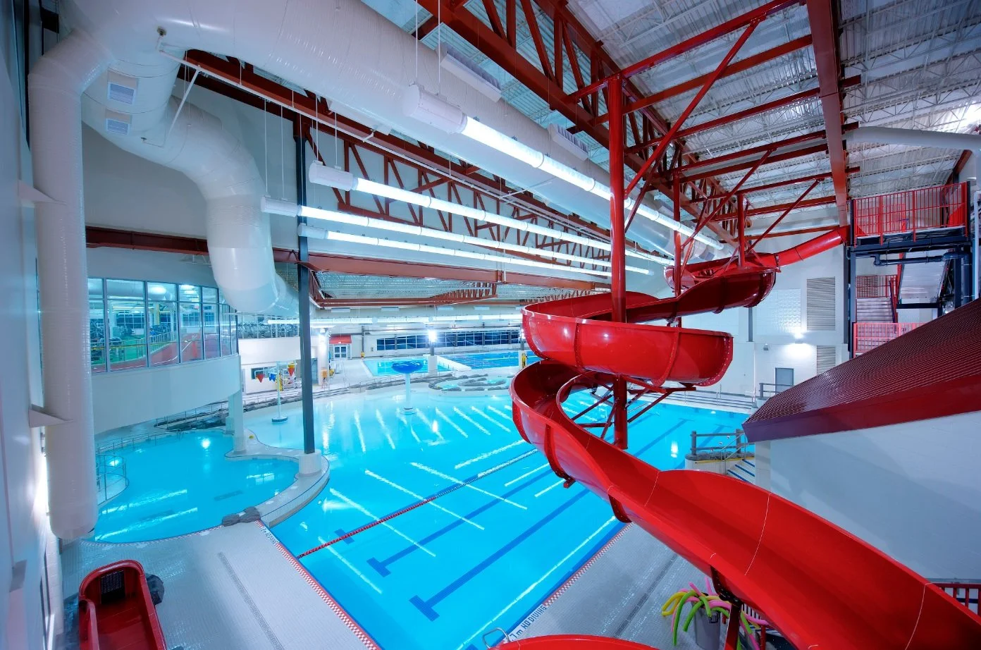 Indoor water park with a large swimming pool, a tall red water slide, and brightly lit interior with white and red structural elements.