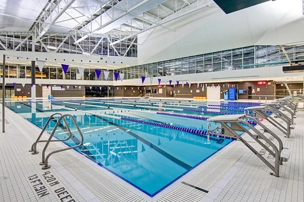 Empty indoor swimming pool with starting blocks and multiple lanes, surrounded by purple banners and a glass ceiling.