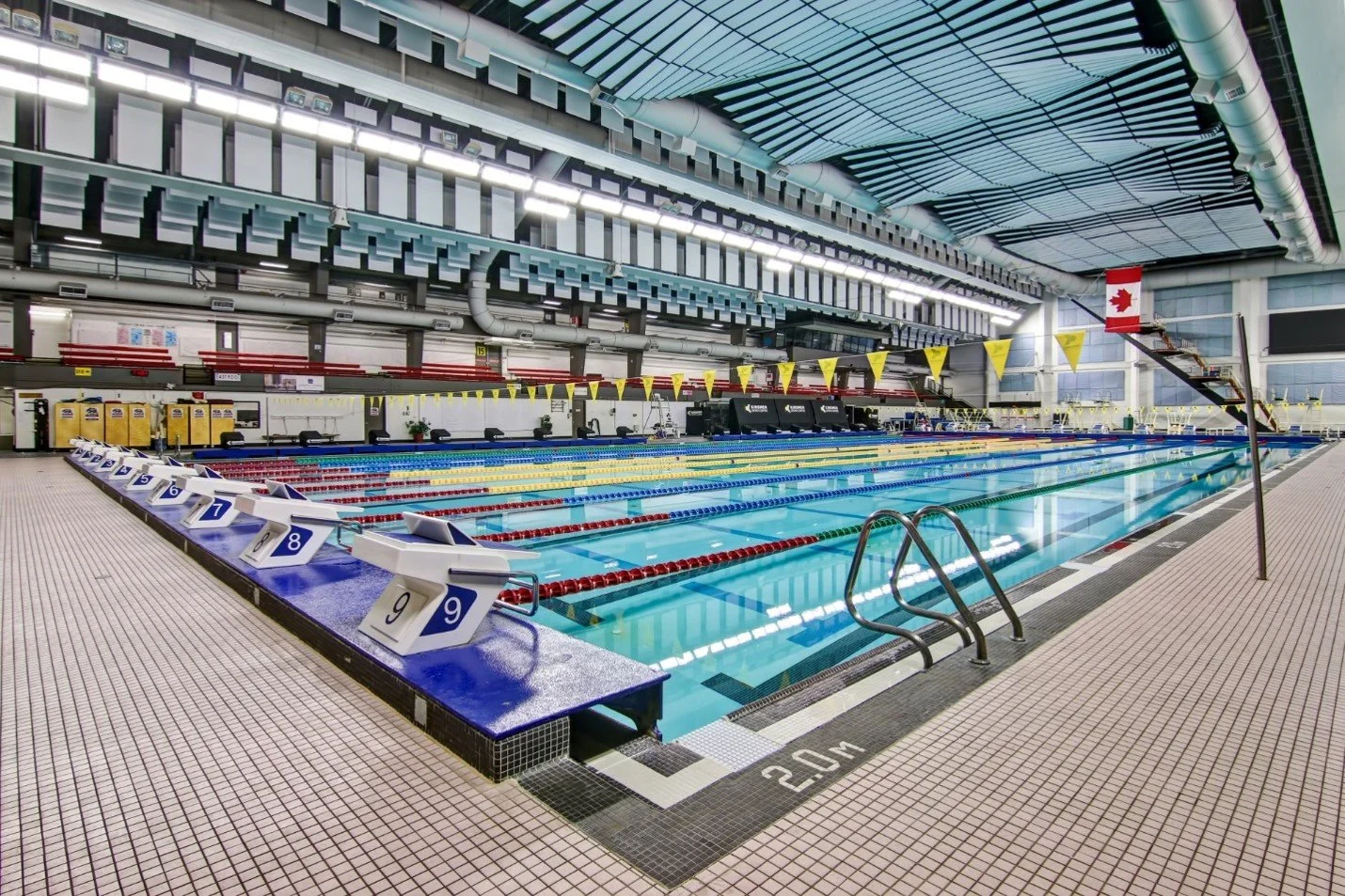 Empty indoor Olympic-sized swimming pool with lane markers and starting blocks, Canadian flag hanging from the ceiling, and bright lighting.