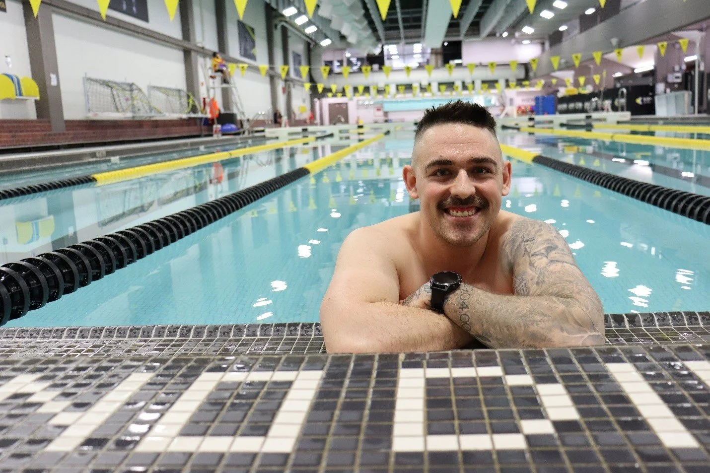 A smiling man with tattoos on his left arm and a watch on his wrist, leaning on the edge of an indoor swimming pool.