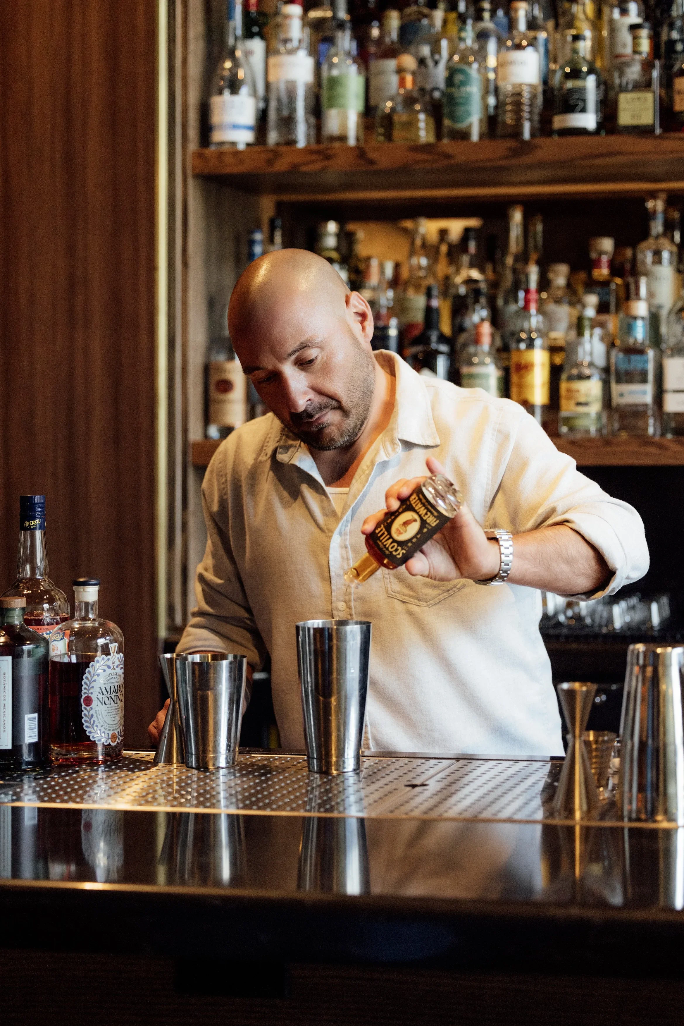 Bald bartender in a beige shirt pouring a liquid into a metal shaker at a bar with various bottles of alcohol on shelves behind him.